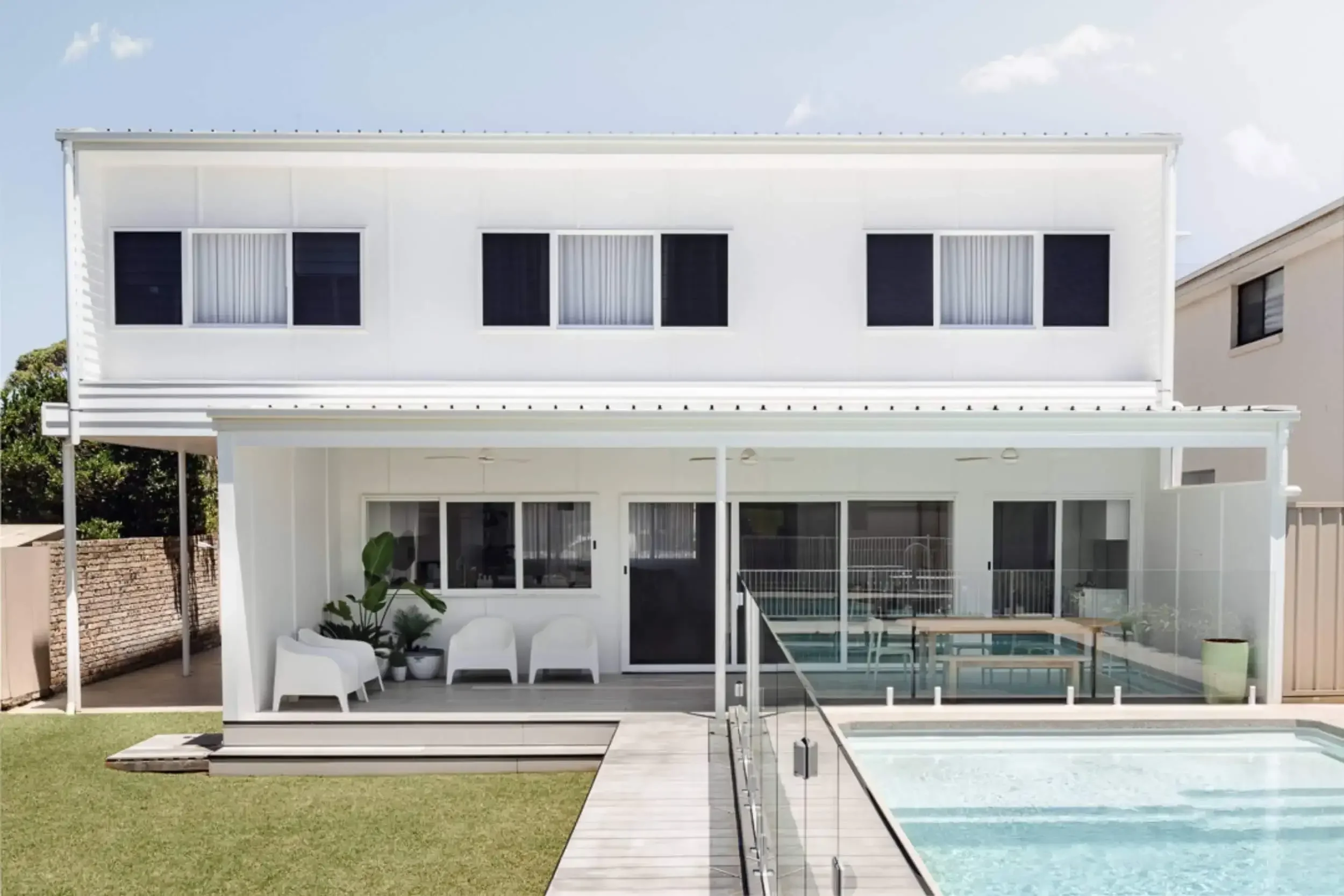 Modern two-story white house with large windows and an outdoor patio. Features a swimming pool, glass fencing, and green lawn. Patio includes white chairs and potted plants under a covered area.