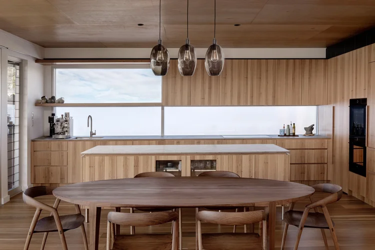 Timber kitchen and long dining table under a timber ceiling at Wombarra Clifftops by CKS Projects
