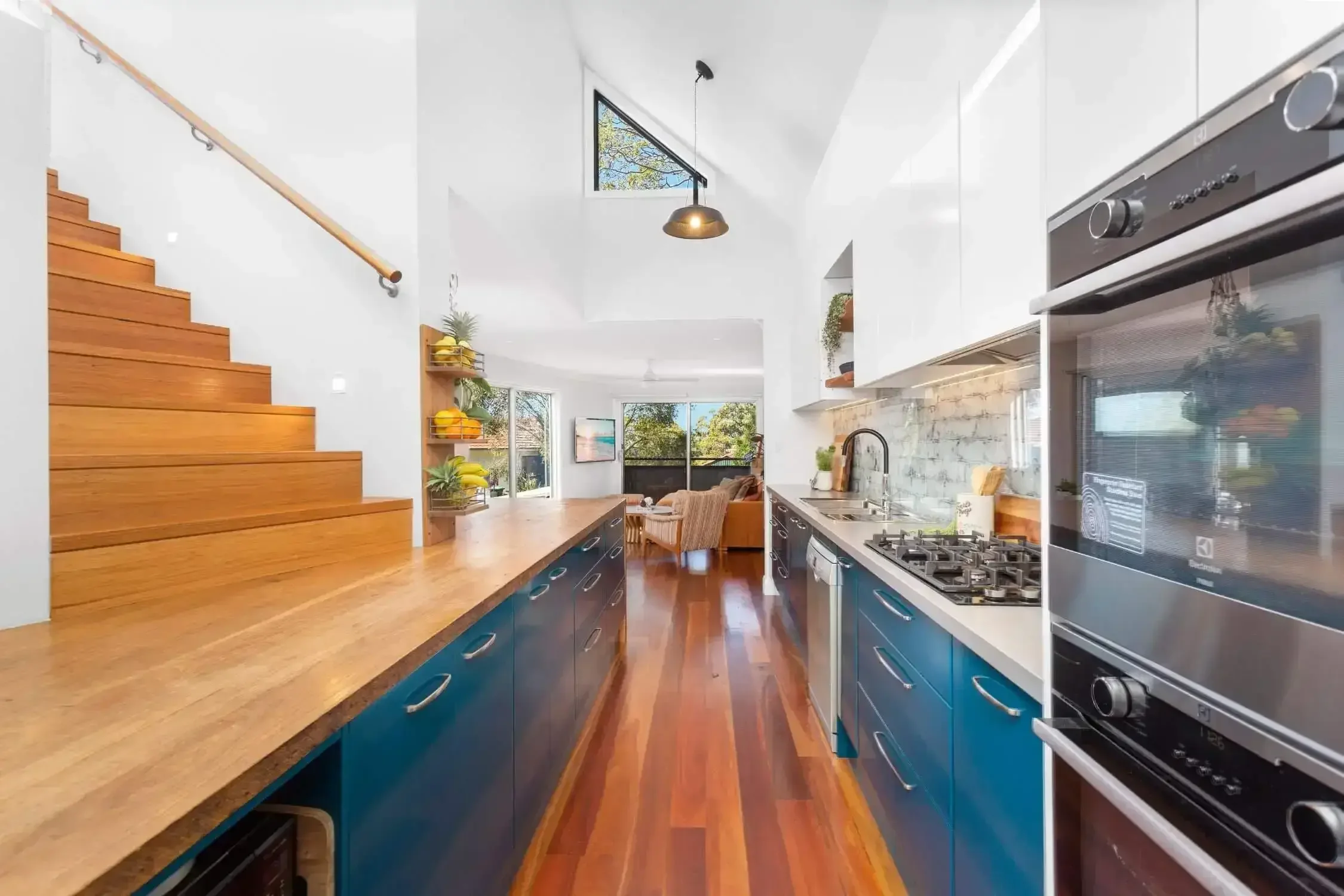 Modern kitchen interior with wooden floors, blue cabinets, and stainless steel appliances. The space features a gas stovetop, built-in oven, light wood countertops, and a decorative fruit display. There's a staircase with wooden steps on the left, le