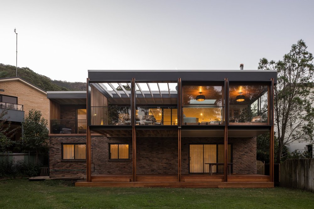 Rear of Elanora Four in Coledale at dusk: a two-level home with timber posts, glazed upper living area and brick lower level above a lawn, by CKS Projects