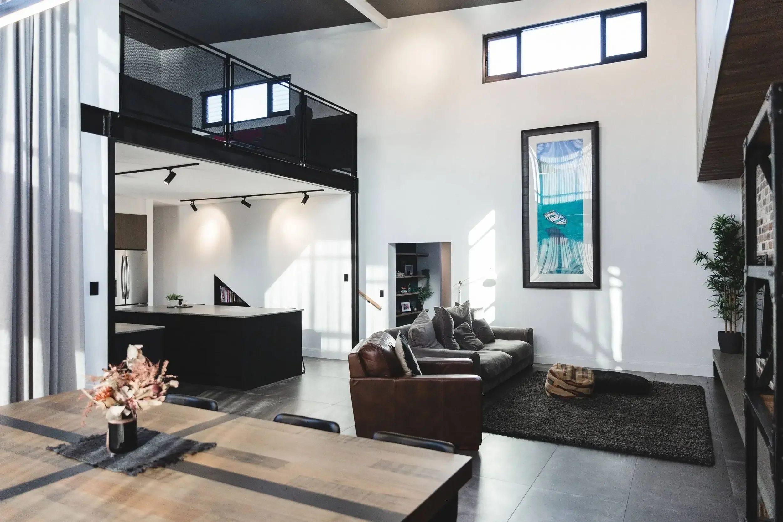 Modern open-plan living room and kitchen with high ceilings, large windows, and contemporary decor. Sofa with cushions, framed artwork, and a small potted plant visible.
