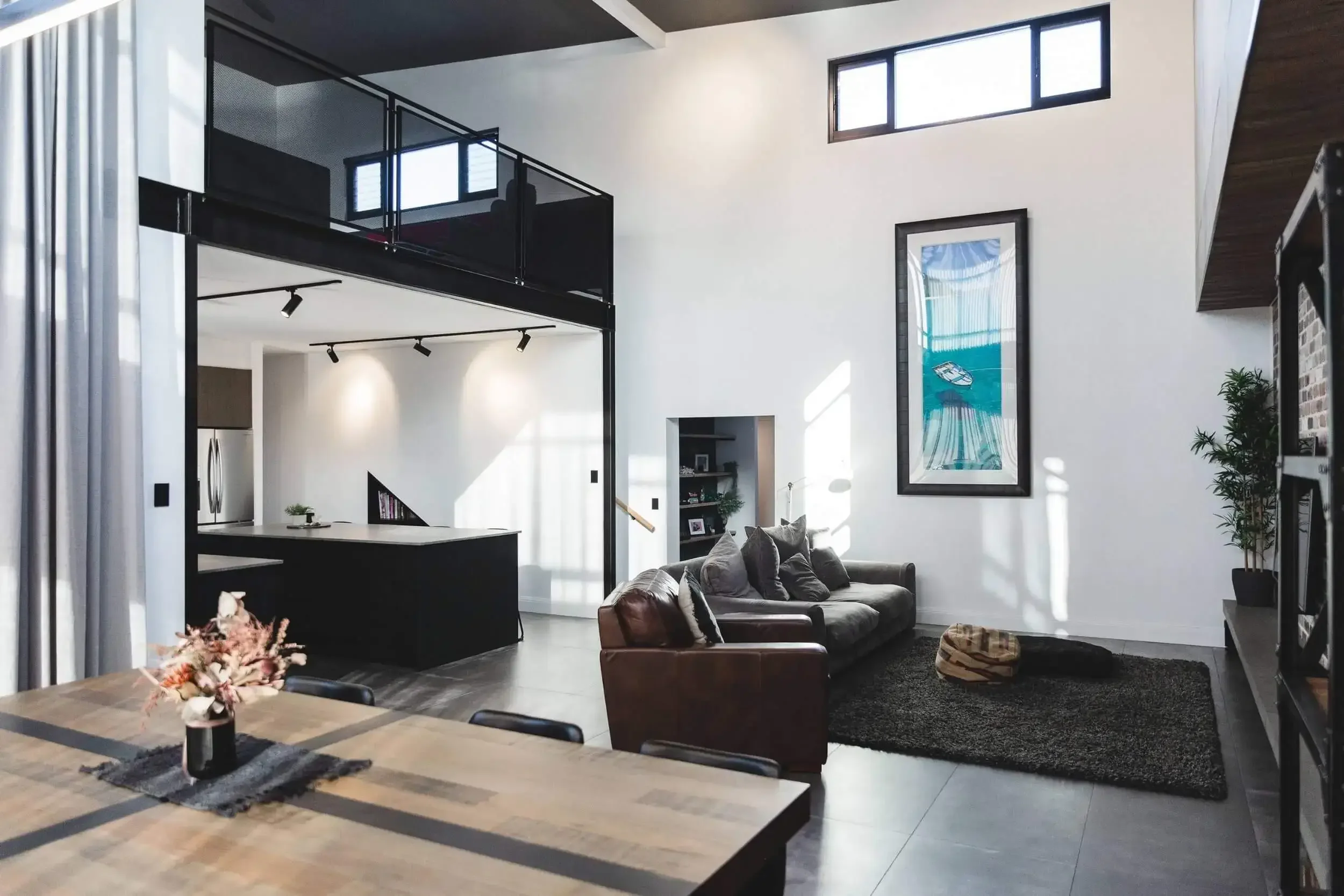 Modern living room with loft, featuring a dining table, sofa, armchair, artwork, potted plant, and a black kitchen island.