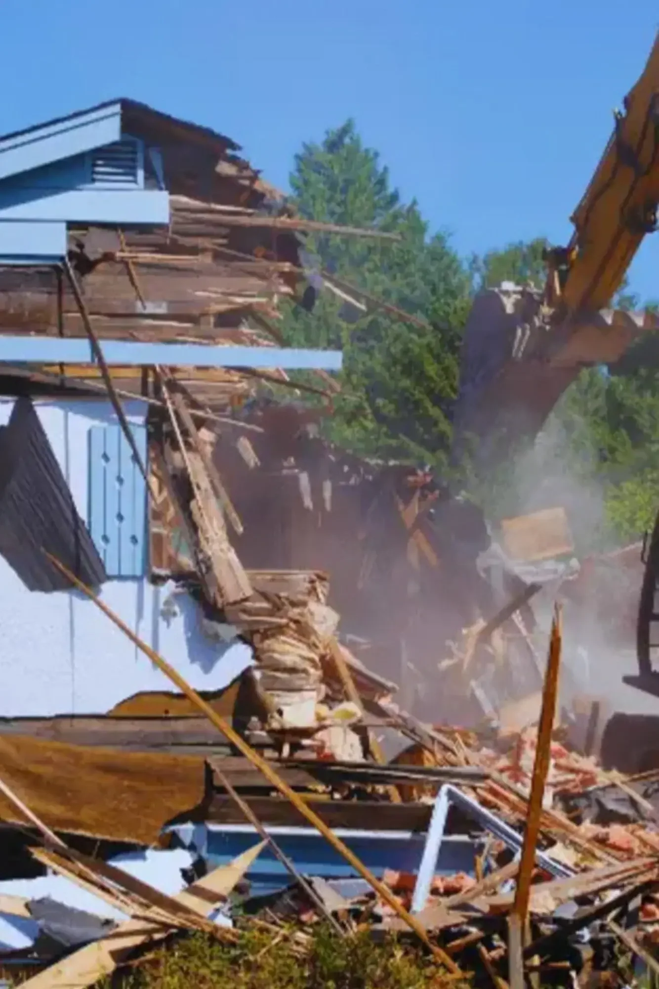 Construction excavator demolishing a house with debris falling, dust rising, and a partially collapsed building in the background.
