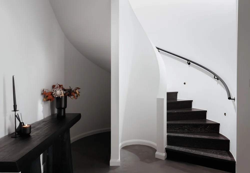 Curved white stairwell with dark timber treads and a black handrail beside a timber console, Huskisson Duplex by CKS Projects