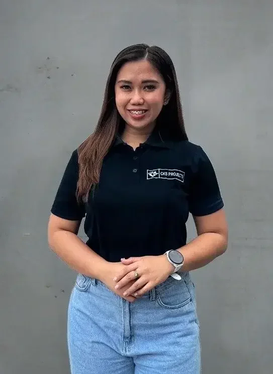 Woman wearing a black polo shirt with "CKS Project" logo and light blue jeans, standing against a gray background, smiling and holding her hands together.