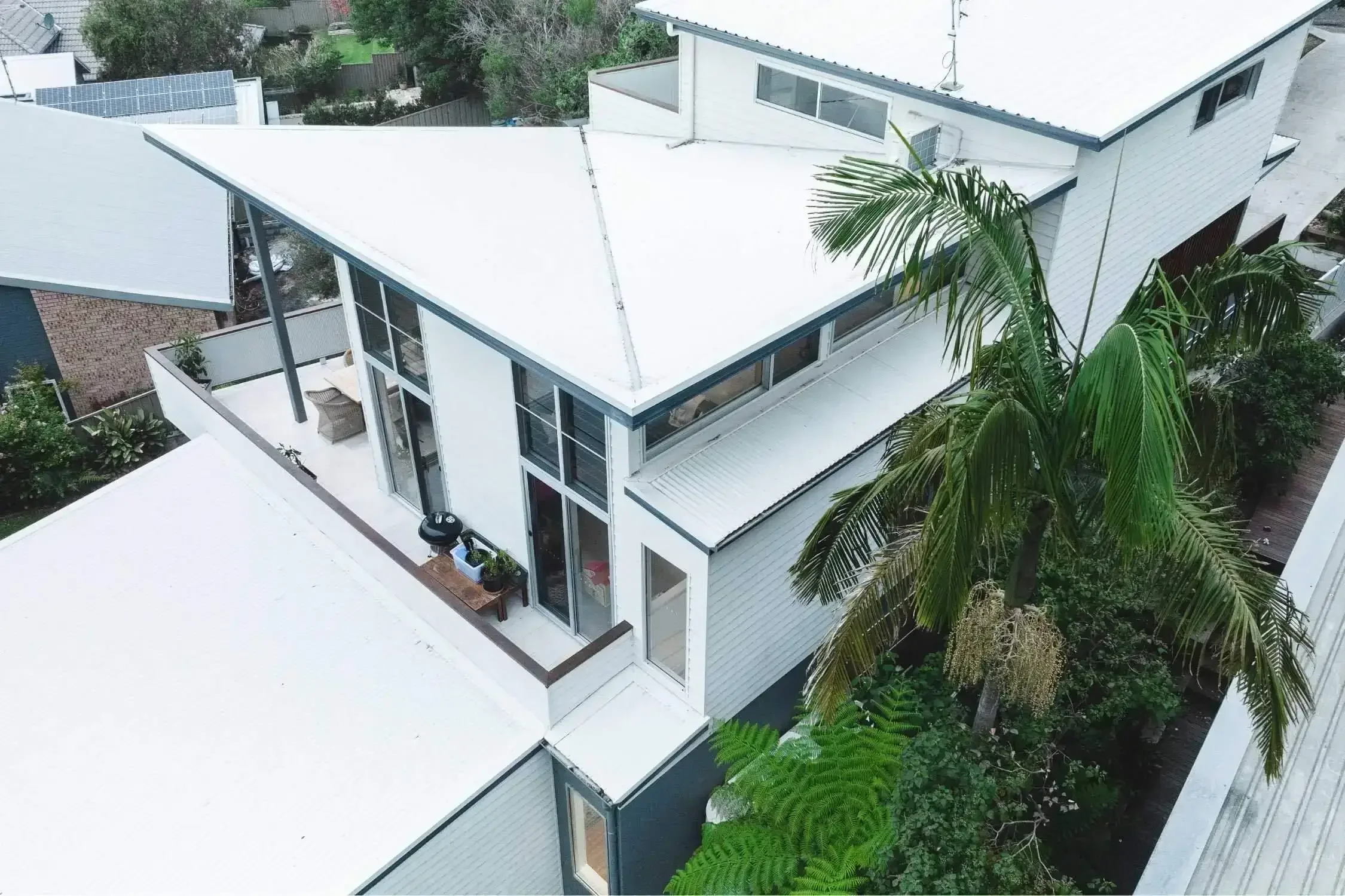 Aerial view of a modern white house with a flat roof and a balcony. The balcony has outdoor furniture and a small grill. The house is surrounded by green trees and vegetation.