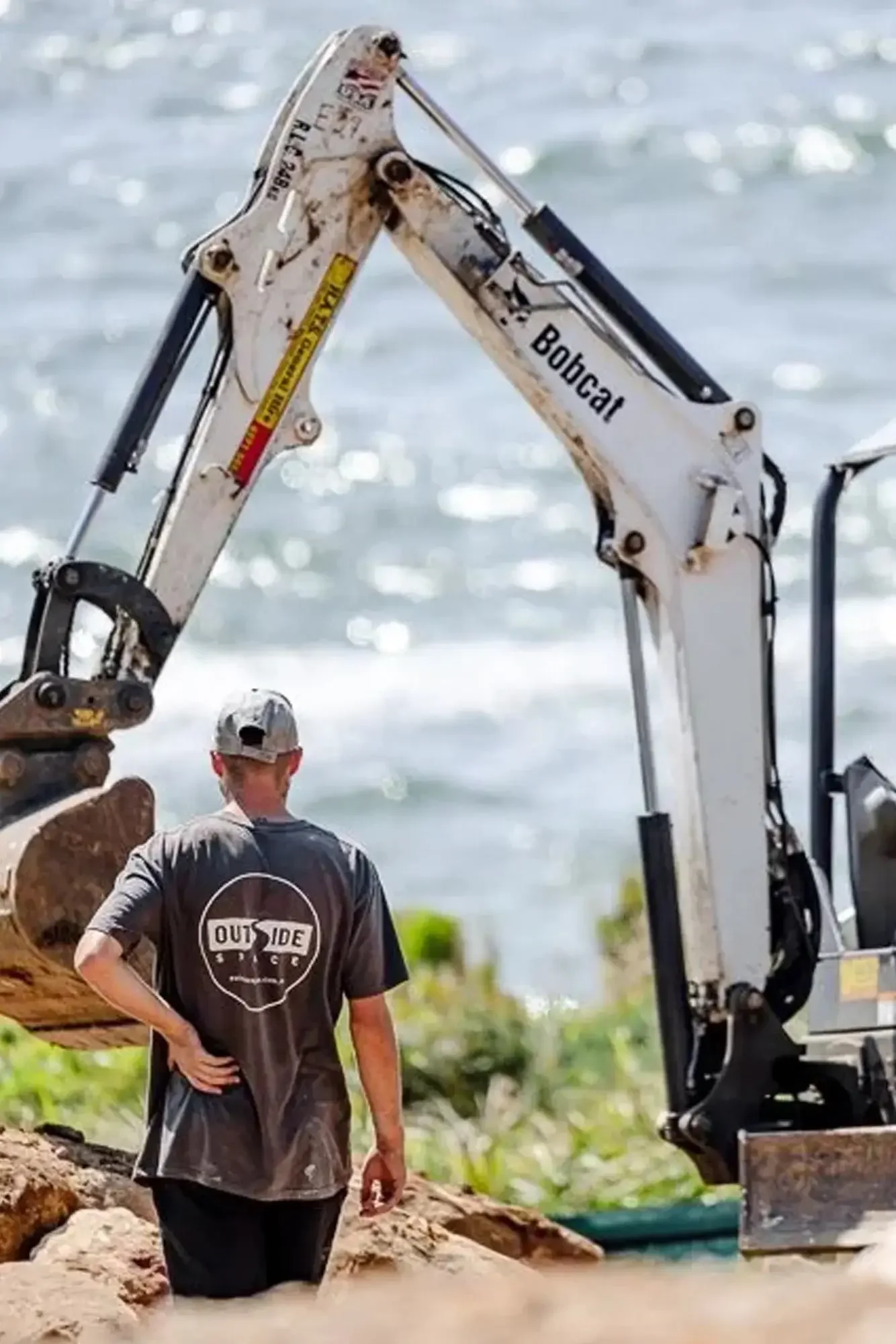 A person wearing a baseball cap and a gray shirt stands next to a large excavator by the water, with the excavator's arm raised. The scene is outdoors with water and greenery in the background.