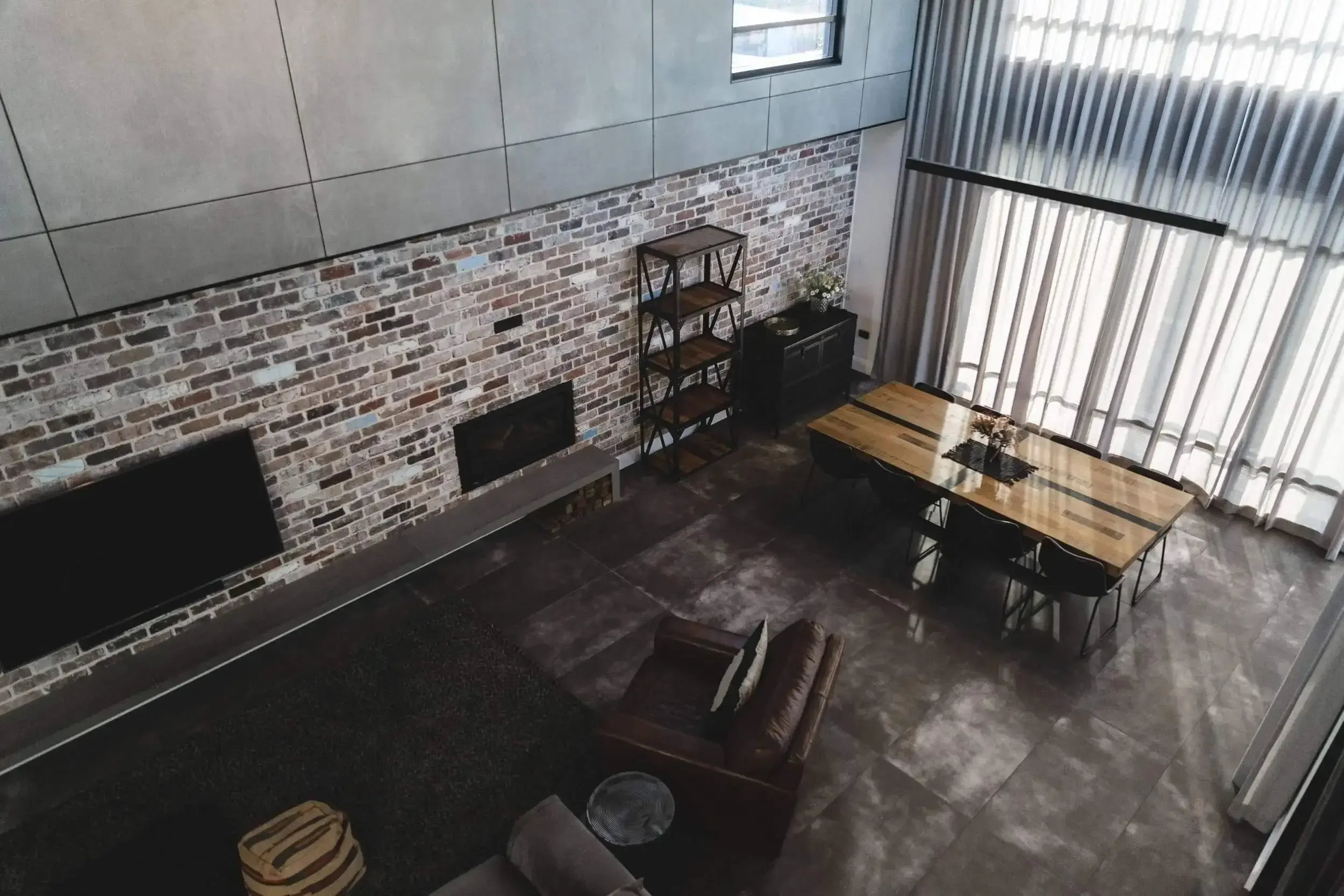Modern living and dining room with grey brick wall, leather armchairs, large wooden dining table, and tall windows with curtains.