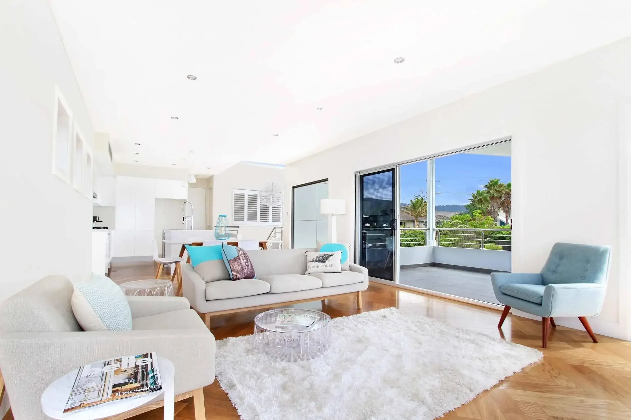 Modern living room with white walls, wood flooring, and sliding glass doors to a balcony. Features a gray sofa, light blue armchair, round glass coffee table, and white rug. Open kitchen area in the background.