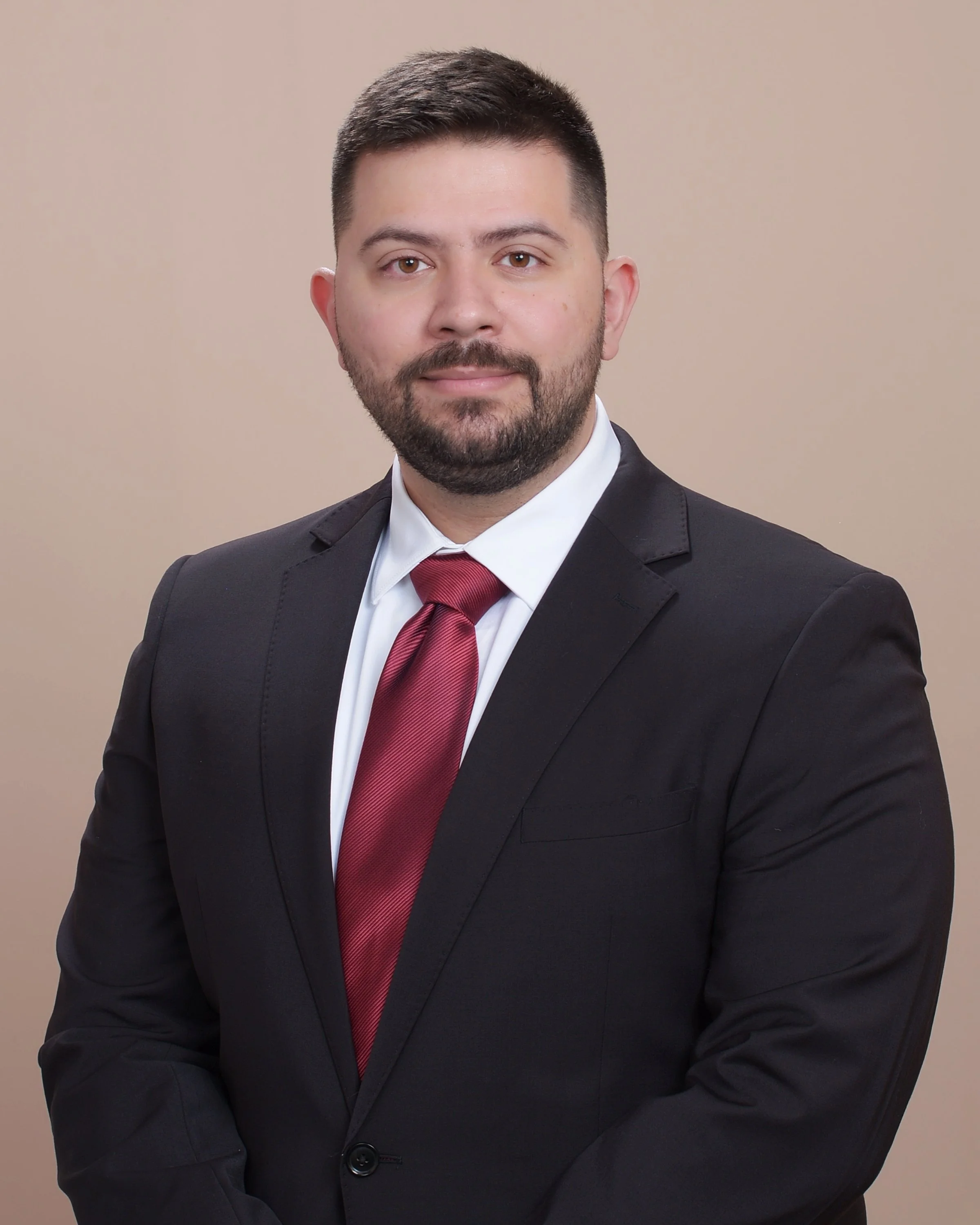 A man in a black suit, white shirt, and red tie posing for a professional portrait against a beige background.