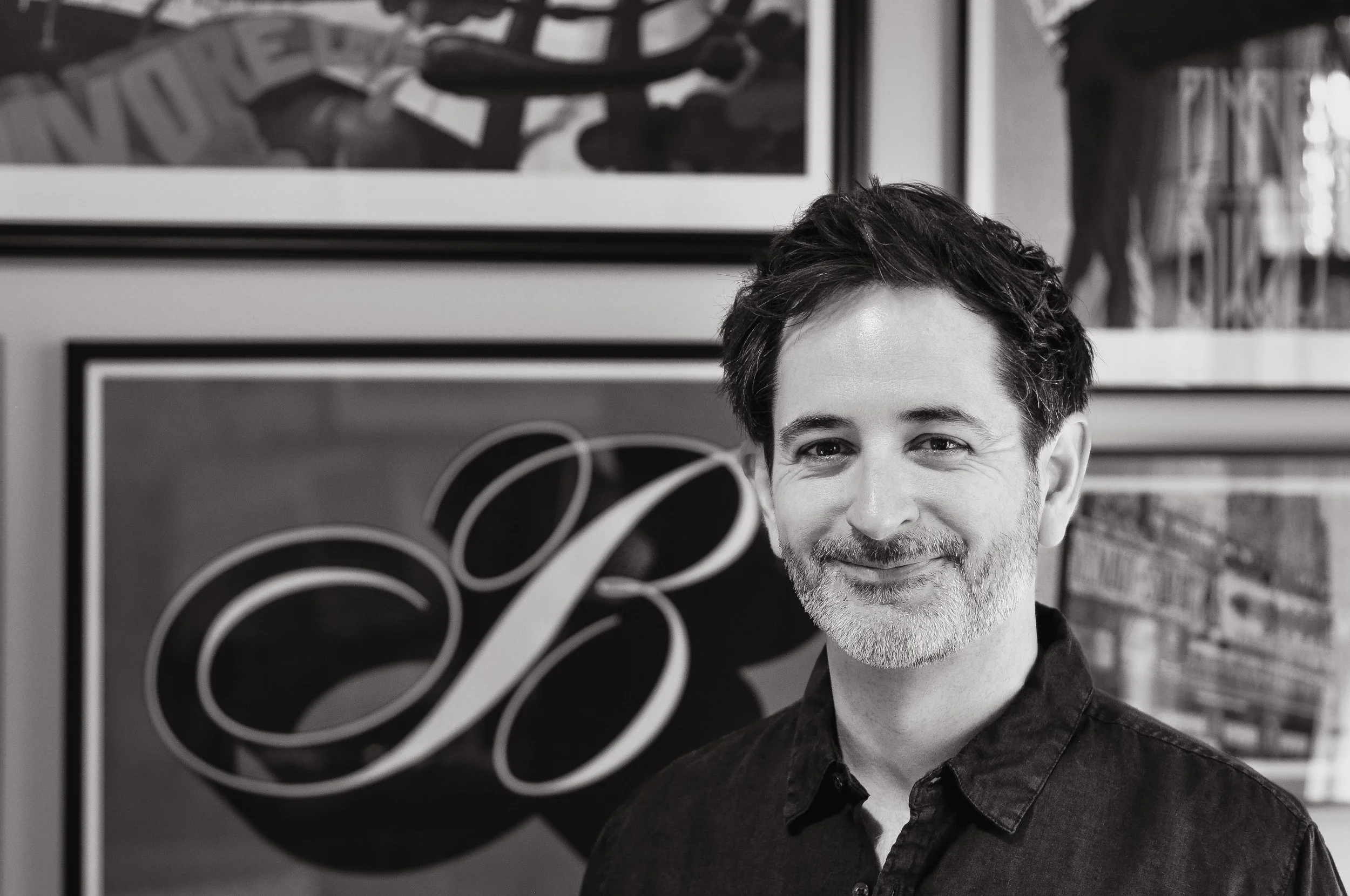 A smiling man with dark hair and a beard, wearing a dark shirt, standing in front of framed artwork including a piece with the letter 'B' in a decorative script.