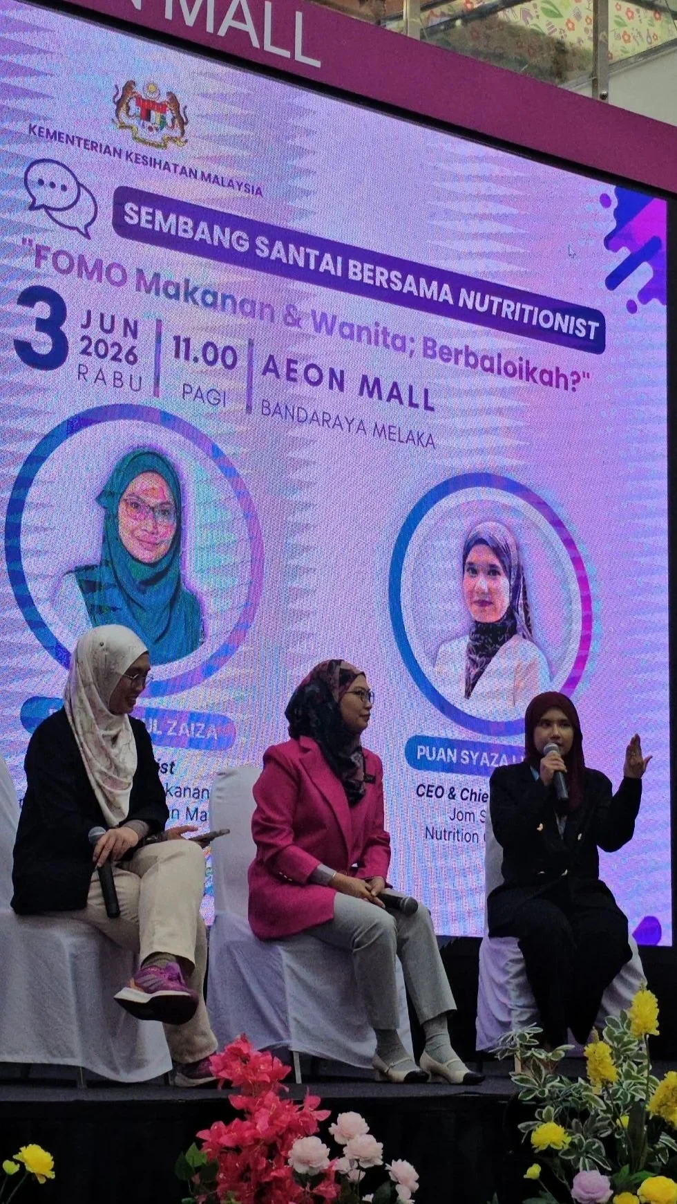 A panel discussion at AEON Mall Bandaraya Melaka about food for women, featuring three women speakers. The background screen displays event details and photos of two speakers, with colorful flowers in the foreground.