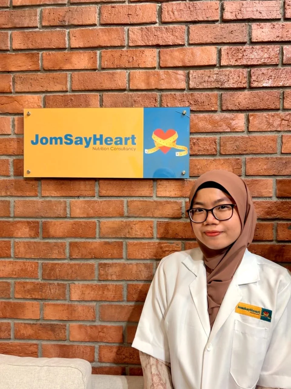A woman in a white medical uniform with a navy blue hijab, smiling, against a white background.