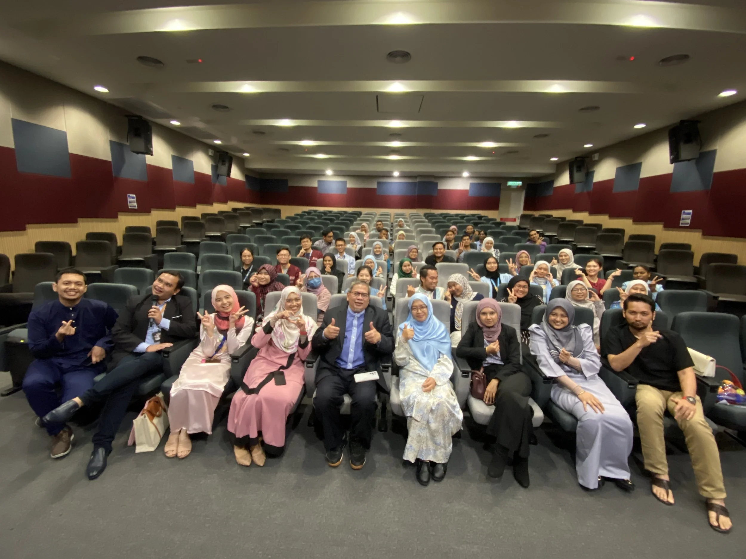 Group of diverse people sitting in an auditorium, smiling, making peace signs, and posing for a group photo.