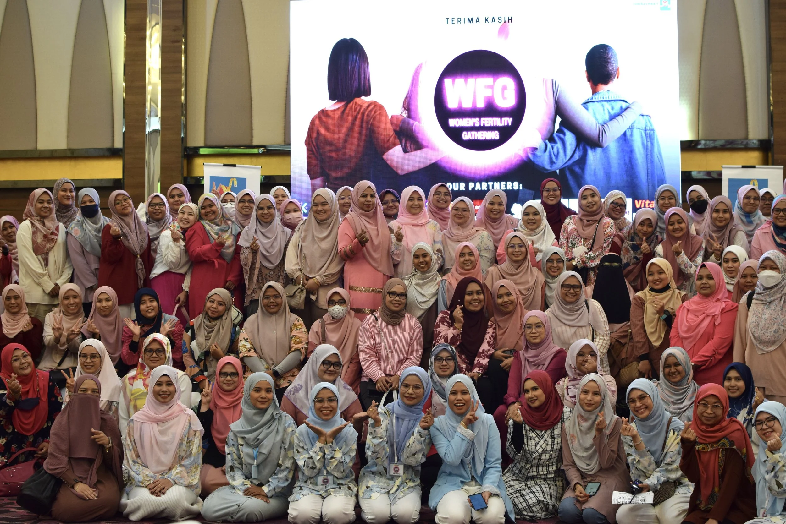A large group of women wearing headscarves and colorful clothing gathered in front of a big screen displaying the Women's Fertility Gathering logo and a message of thanks.