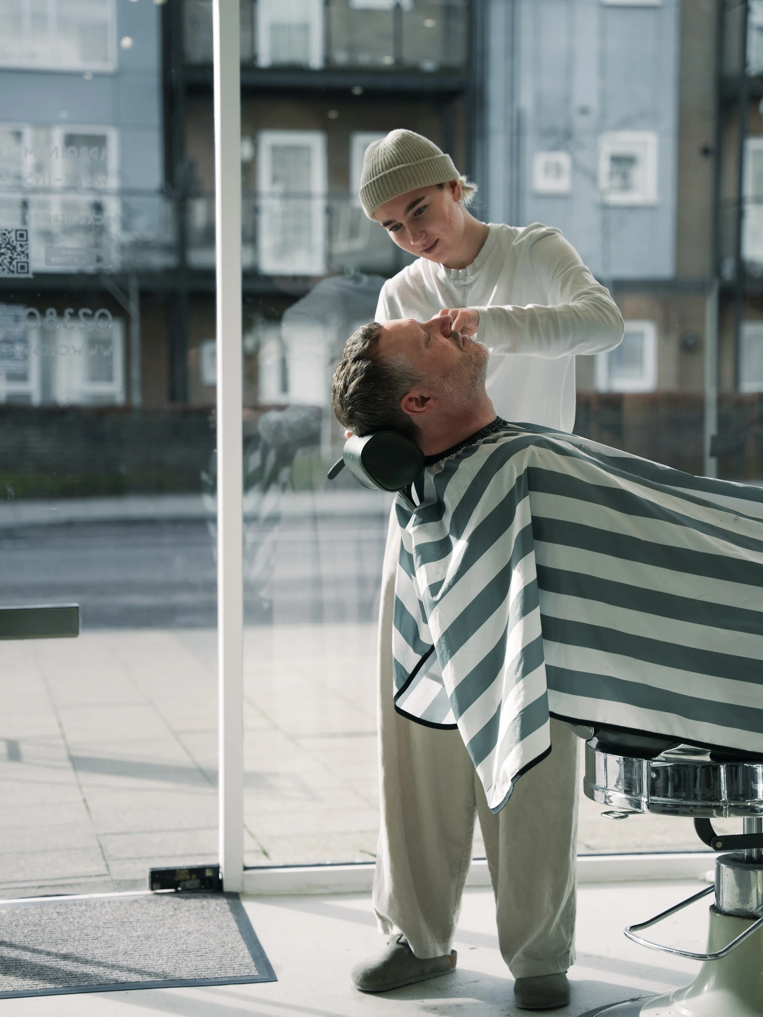 Charlotte Webb is a barber who works at Cresswell Barber Co. In this photography she is smiling, wearing a beanie, tracksuit bottoms and is cutting a man's hair who has a green and white striped cape on.