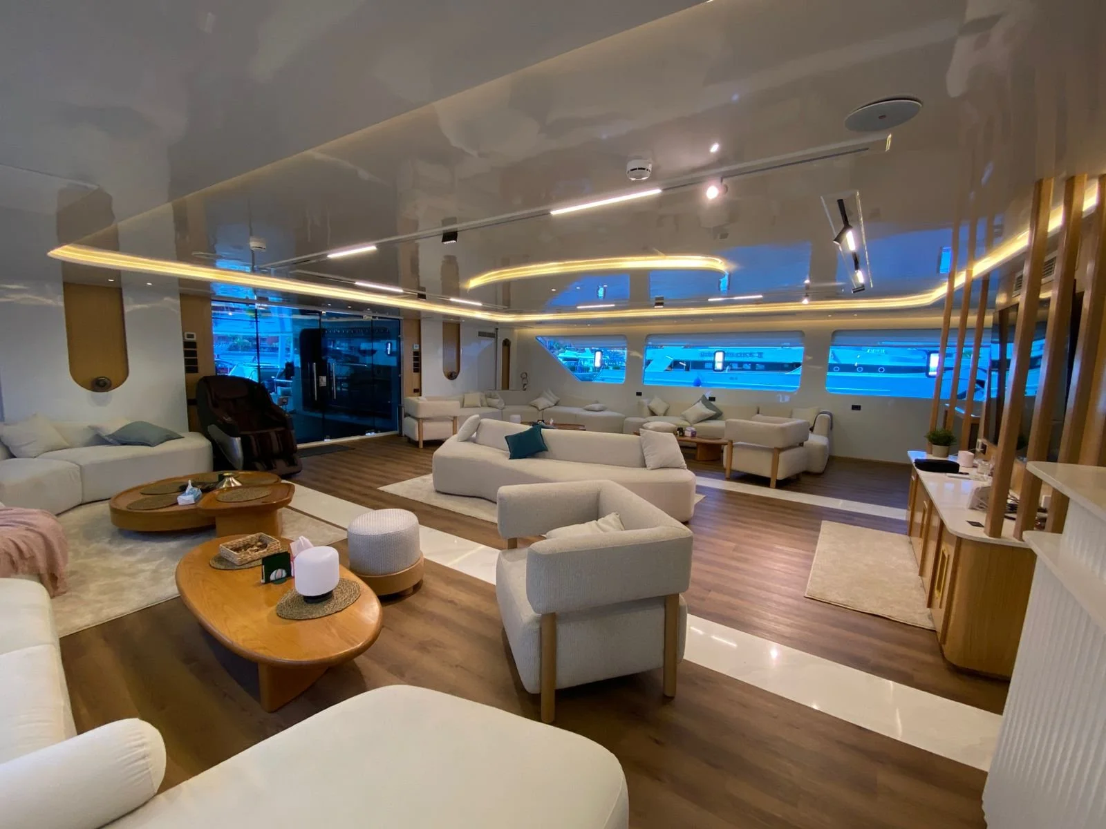 Luxury yacht interior with white couches, wooden coffee tables, a massage chair, and large windows showing a marina with boats at blue dusk.