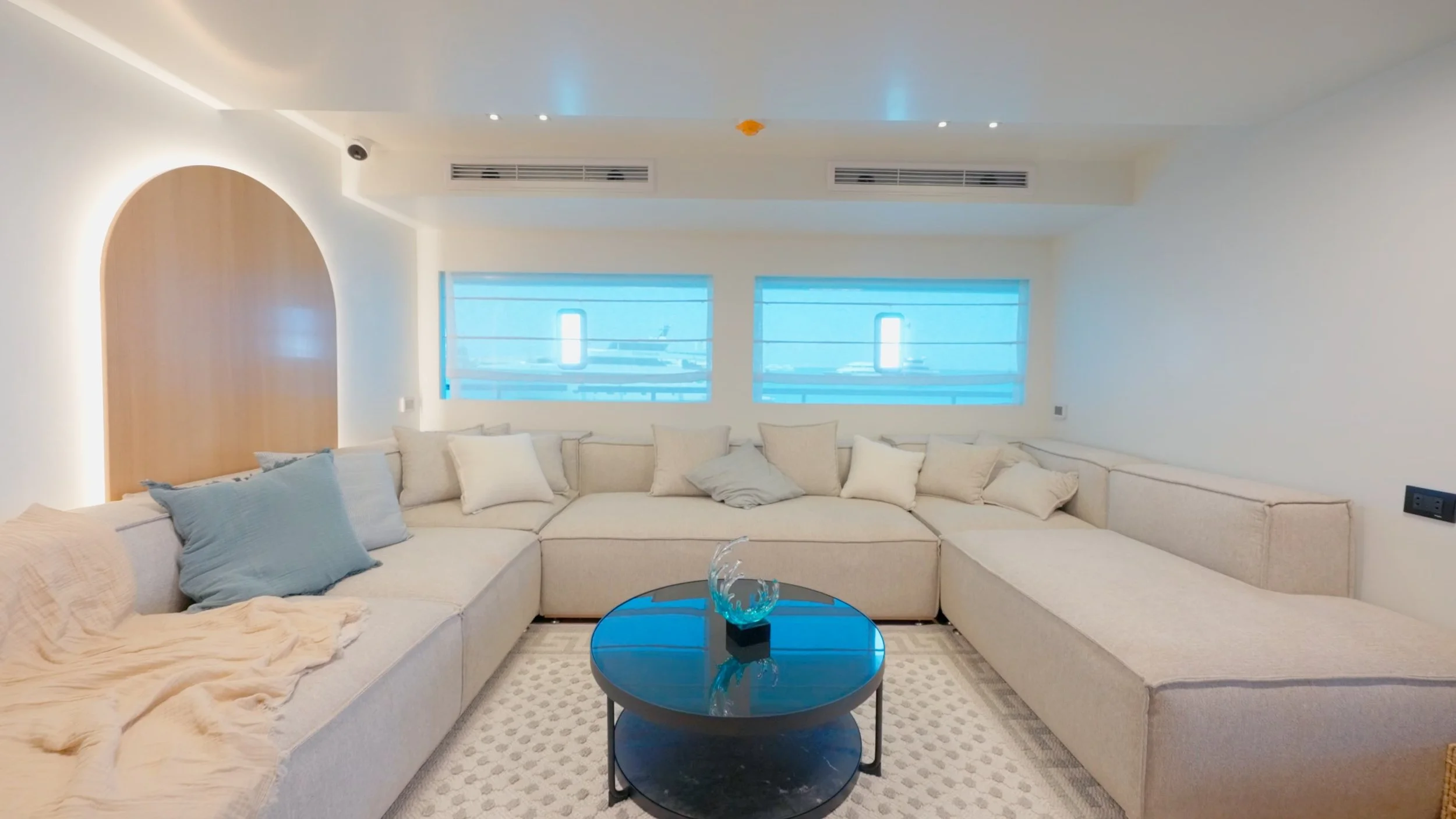 Living room with a large off-white sectional sofa, blue and beige pillows, a black round coffee table with a glass sculpture, and three windows with blue shades.
