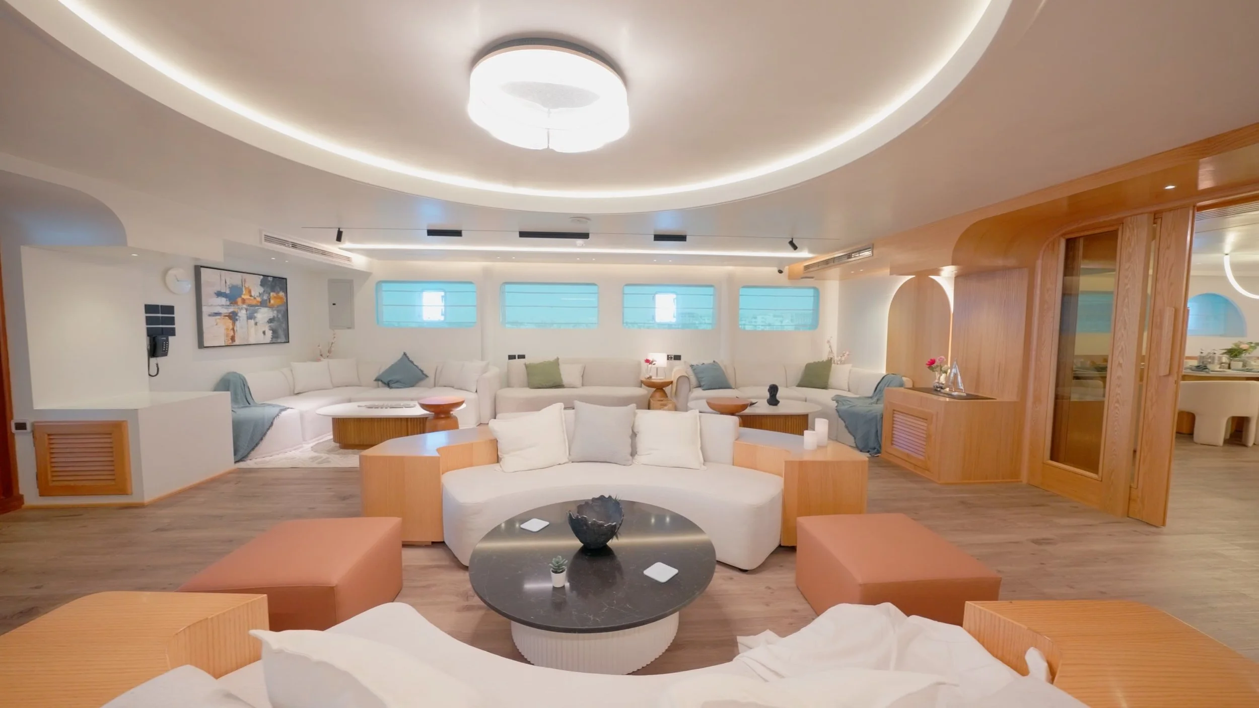 A spacious, modern living room with a circular arrangement of white sofas, a round black coffee table with decorative items, and large windows with blue screens. The room features wooden accents and ceiling lighting.