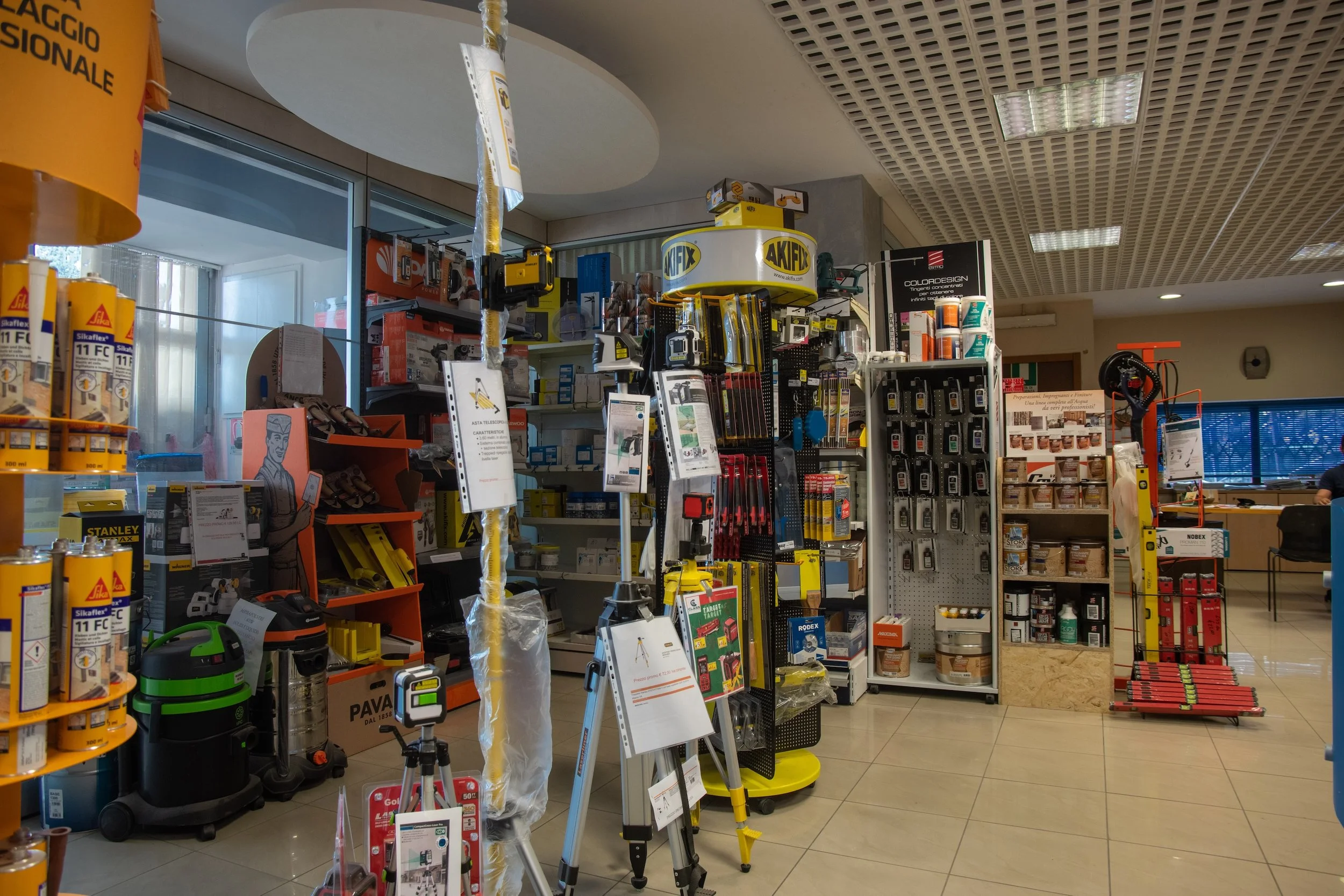 Innenansicht eines Baumarkts mit Werkzeugen, Sicherheitsausrüstung und Baustoffen. Regale voller Produkte und Werkzeuge auf Fliesenboden.