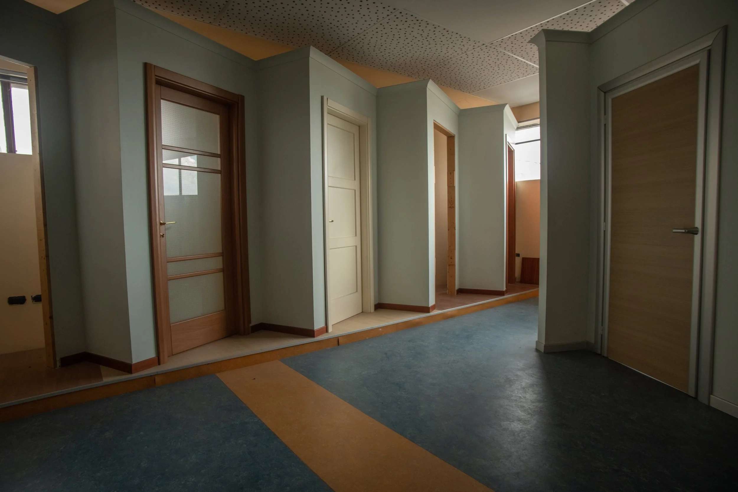 Interno di un edificio con porte in legno e pareti verdi, pavimento in piastrelle di vari colori.
