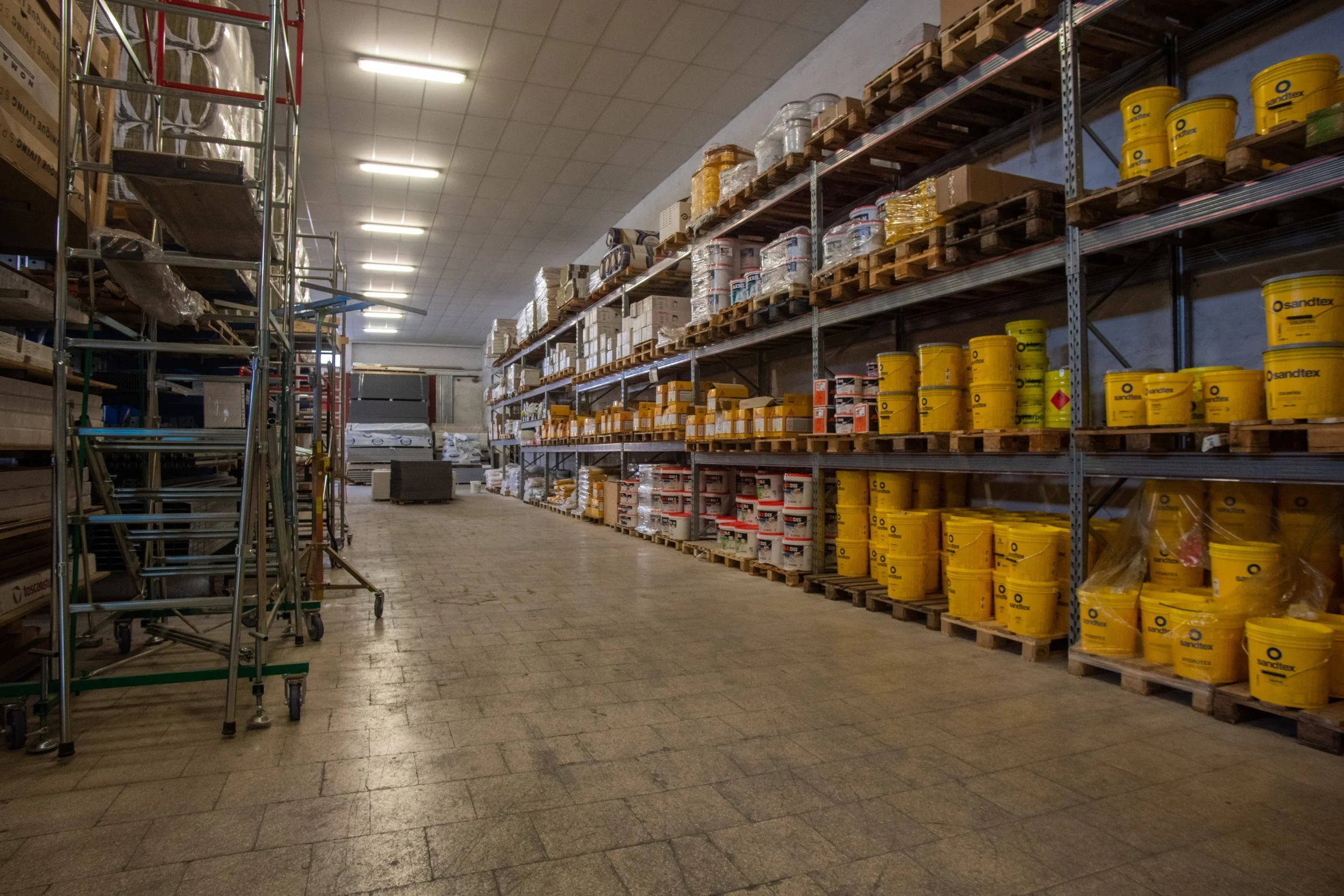Magazzino con scaffalature di prodotti per edilizia, casse di vernici gialle e integrazioni, su pallet di legno, scaffale metallico e pavimento in mattonelle