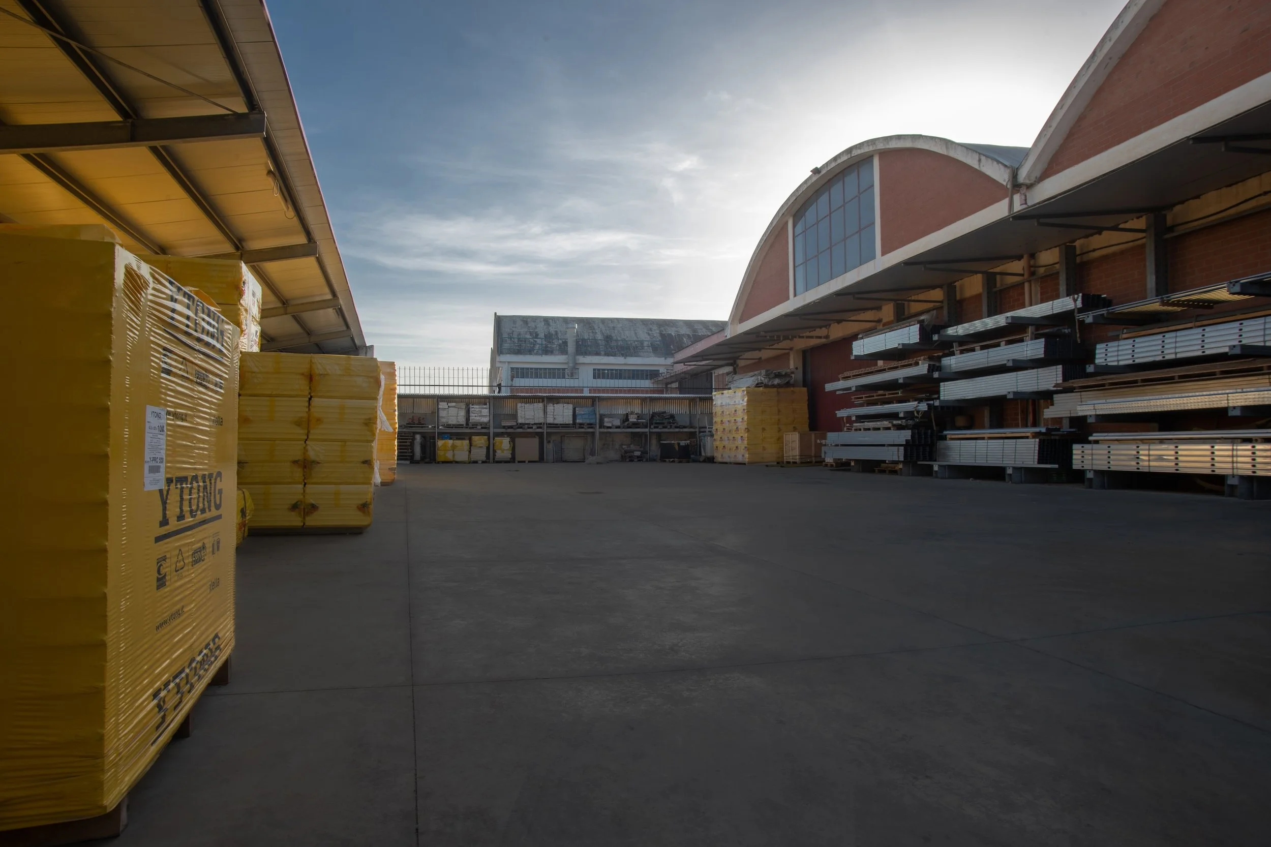 Scaffali di materiali edili sotto una tettoia in un cortile industriale, con un cielo nuvoloso e il sole basso all'orizzonte.
