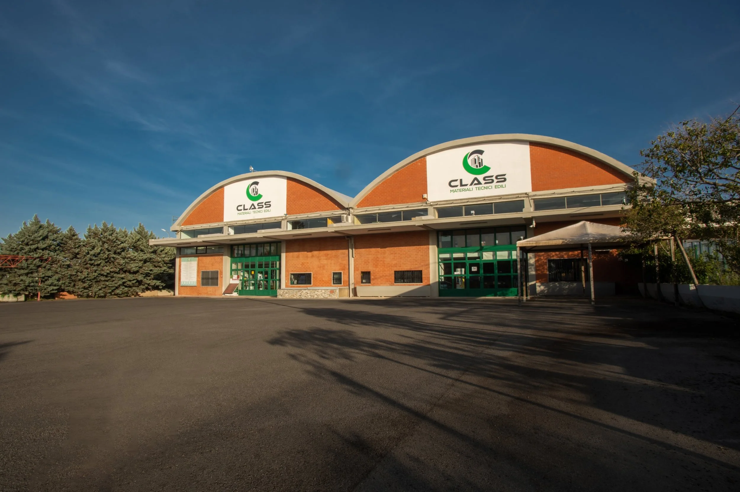 Edificio con segnaletica che legge 'CLASS Materiali Tecnici Edili', pareti di mattoni rossi, porte e finestre verdi, sotto un cielo sereno