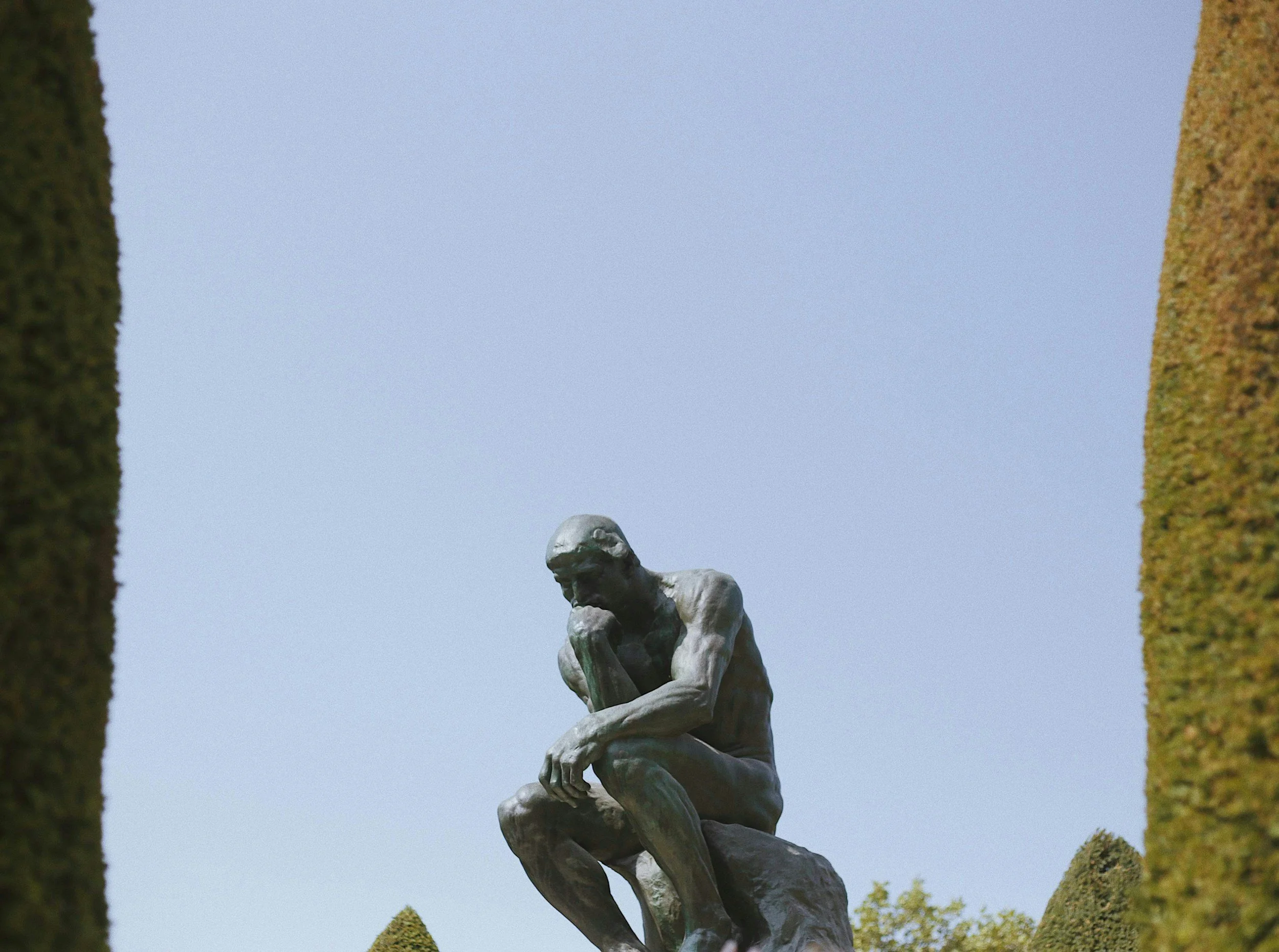 A bronze statue of a man sitting on a large rock, with one hand resting on his knee and his head bowed. The statue is located outdoors between two tall, narrow trees with a clear blue sky in the background.