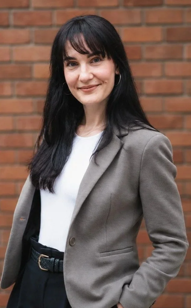 A woman with long black hair and hoop earrings, smiling, standing outdoors in front of a brick wall, wearing a gray blazer over a white top.
