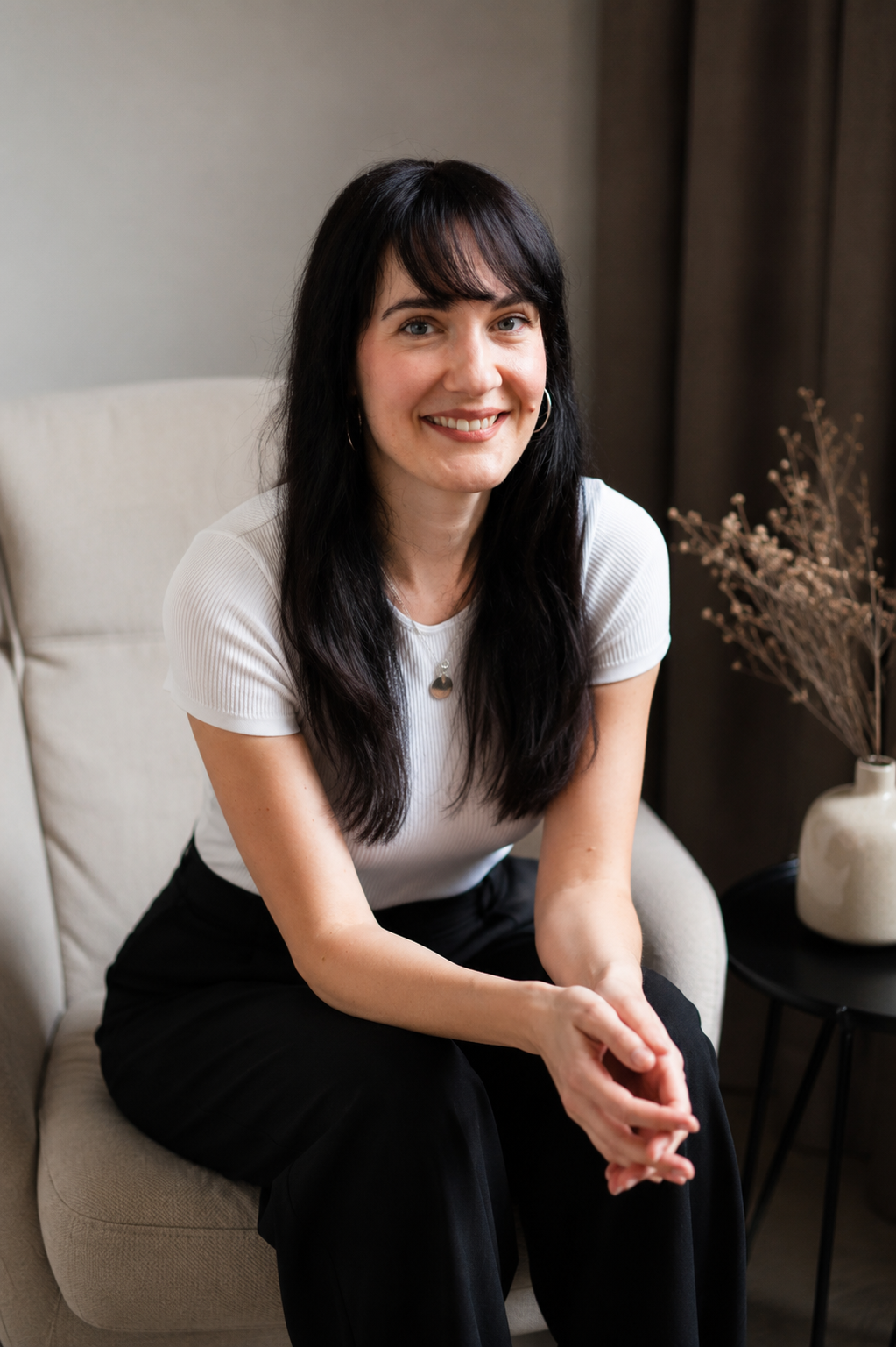A woman with long dark hair and hoop earrings, smiling, sitting on a beige chair in a room with dark curtains and a side table with a white vase and dried flowers.