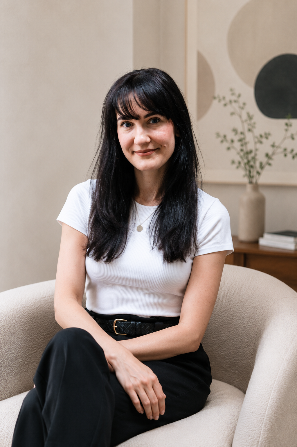 A woman with long dark hair and bangs sitting on a beige couch in a room with neutral-colored walls and artwork, wearing a white t-shirt and black pants, smiling at the camera.