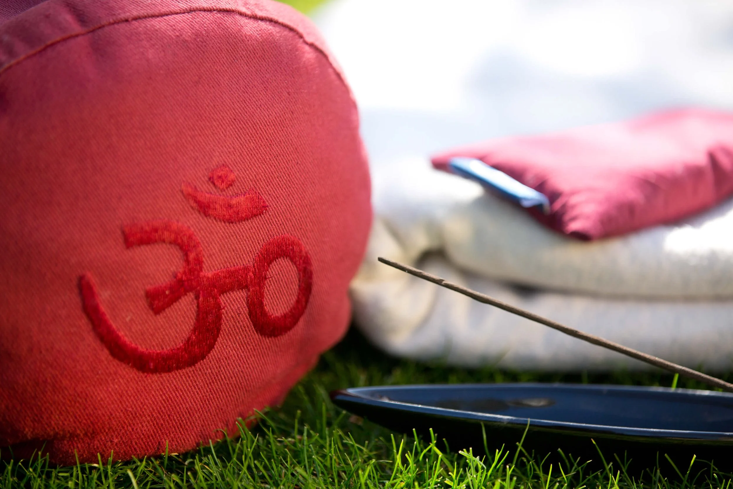 A red fabric yoga bolster with a stitched om symbol, resting on grass, with a white cloth, a pink pillow, and a black meditation bell nearby. at move well studio wetherby