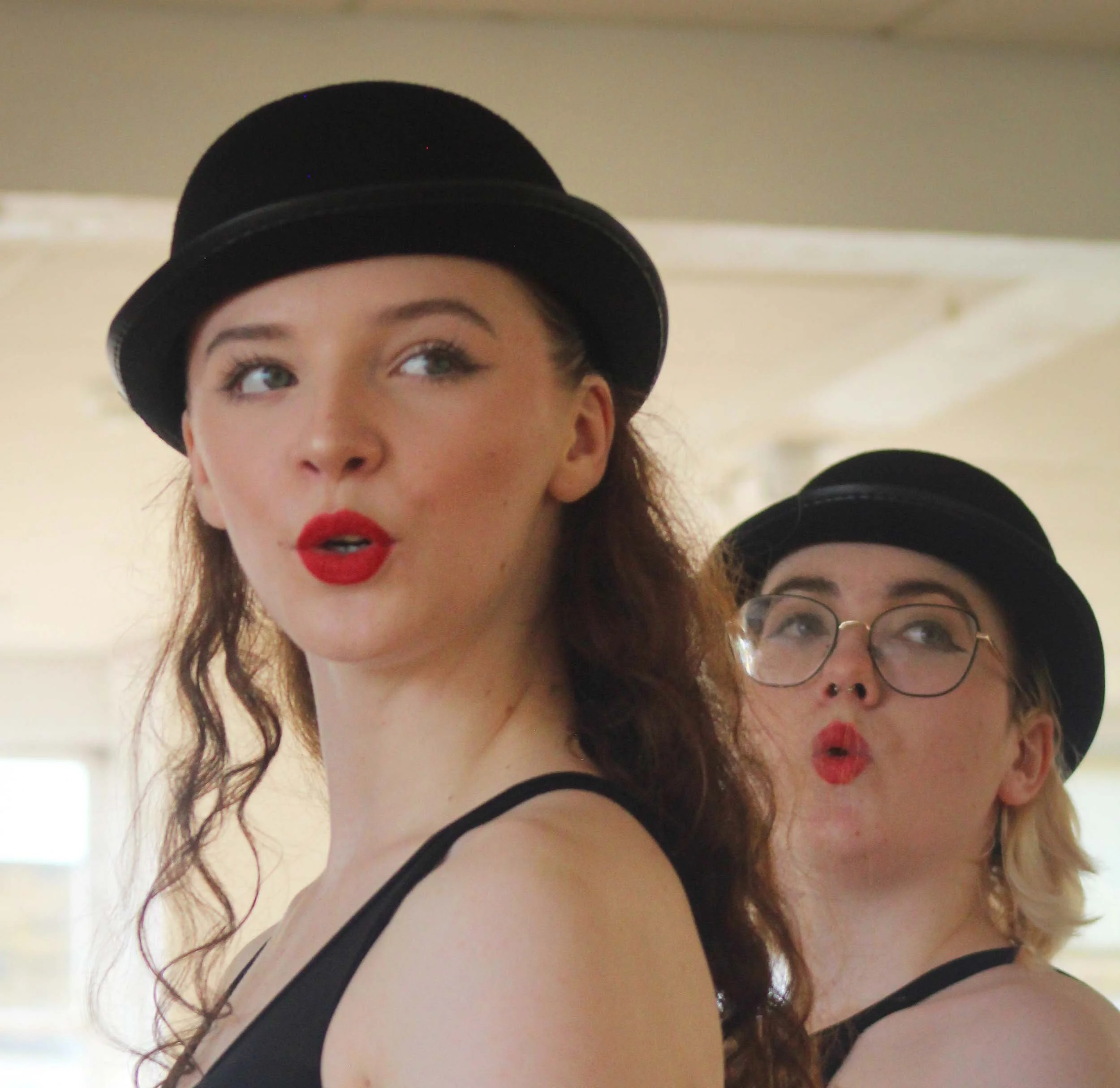 Two dancers in bowler hats and red lipstick, close-up in a bright studio, one in focus and one behind.