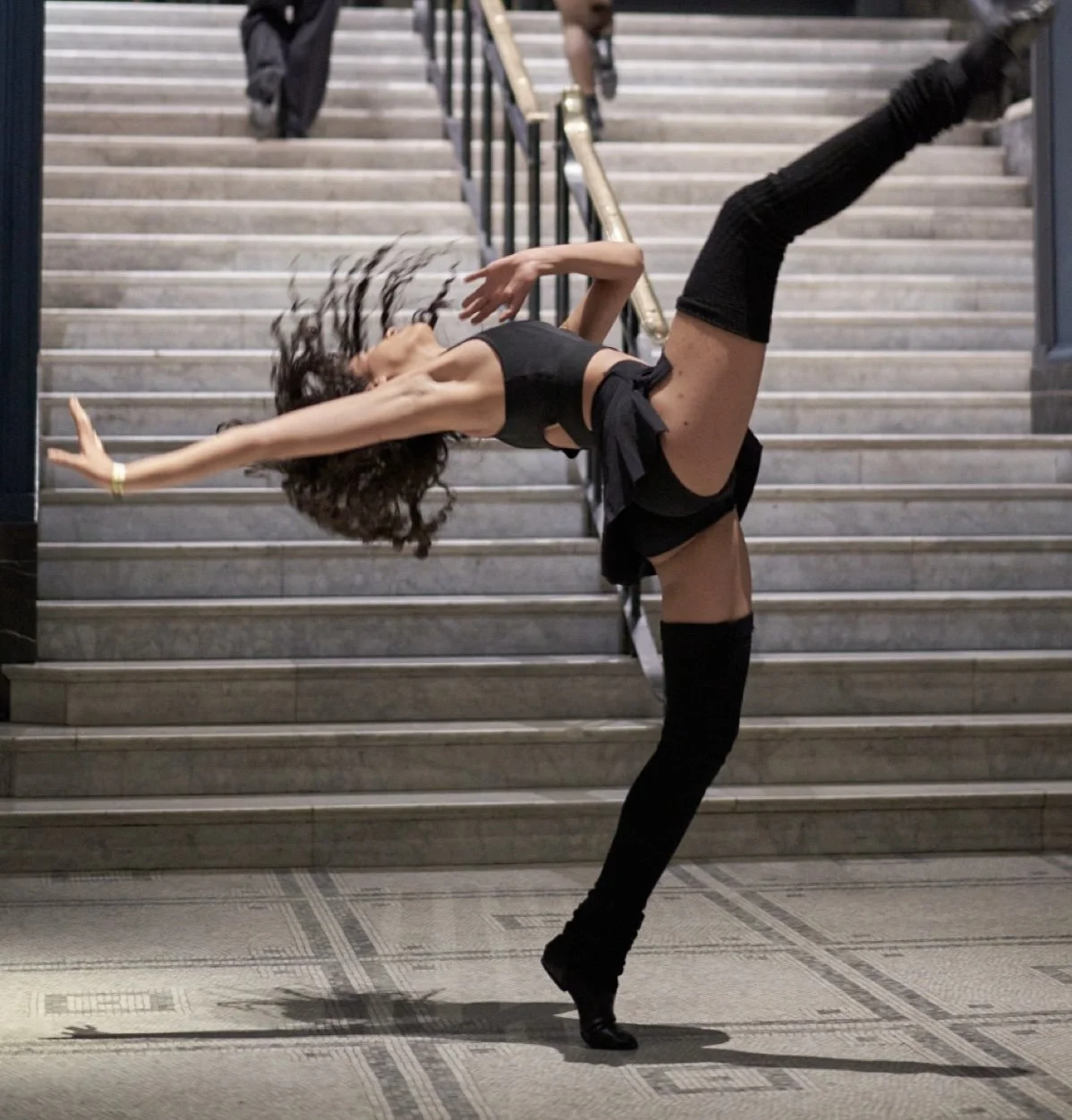 Dancer arches backwards with one leg raised high on a grand marble staircase, hair in motion, wearing all black