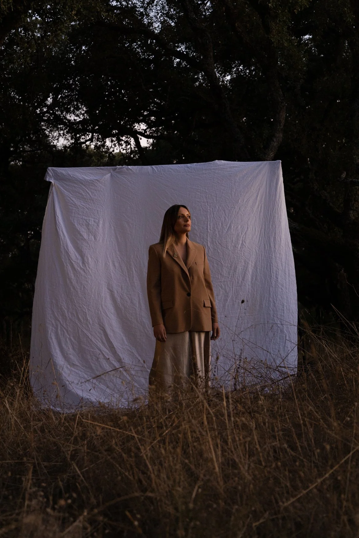 Mulher de terno bege diante de um pano branco ao ar livre, com árvores ao fundo.