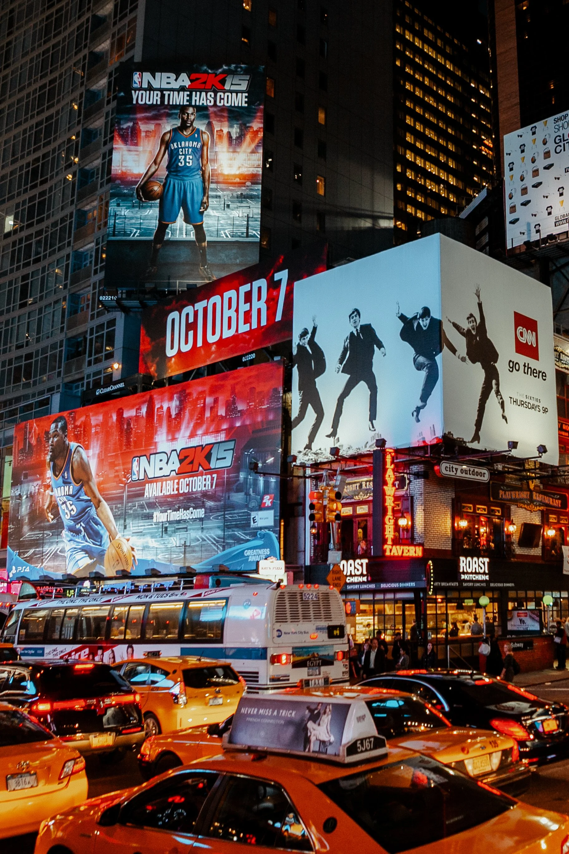 Times Square à noite, com outdoors iluminados exibindo anúncios do NBA 2K15 e outras propagandas, carros e táxis ocupando a rua.