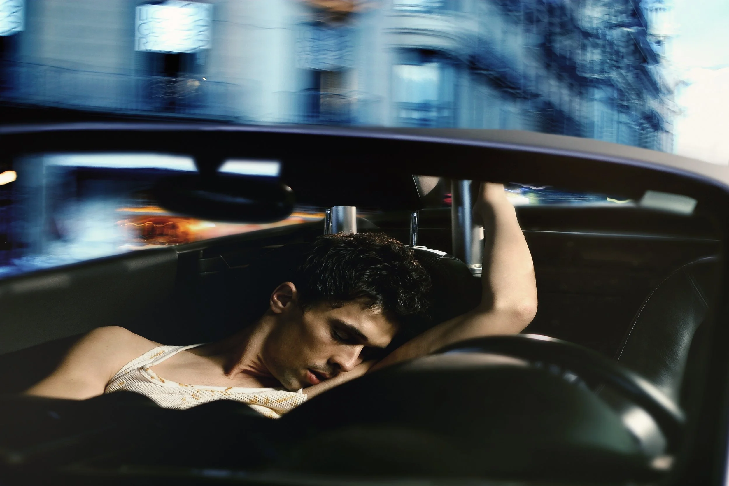 Homem deitado em um carro em movimento, com fundo desfocado de prédios.