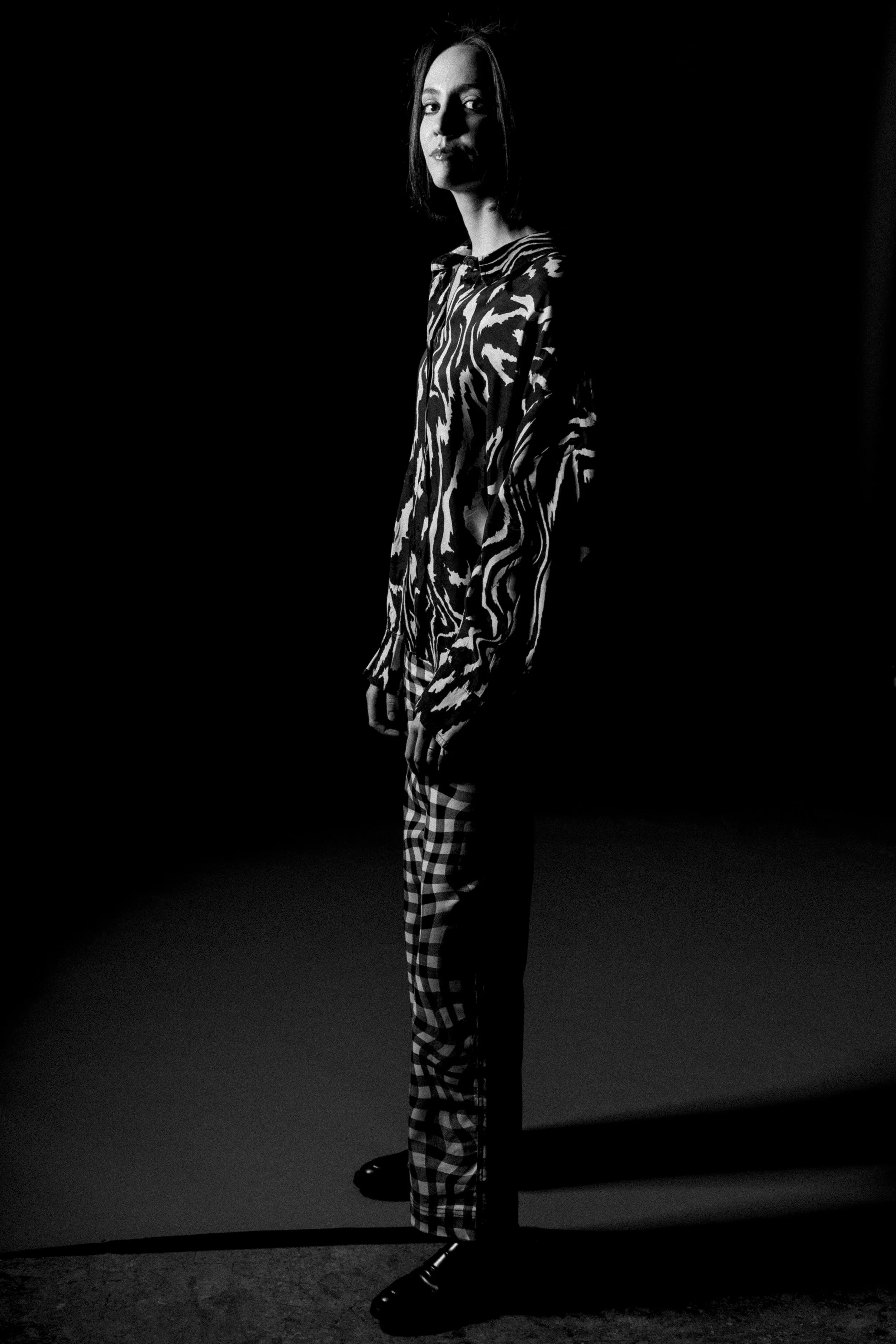Pessoa em pé, vestindo roupa com estampa zebrada e calça xadrez, em um ambiente mal iluminado em preto e branco.