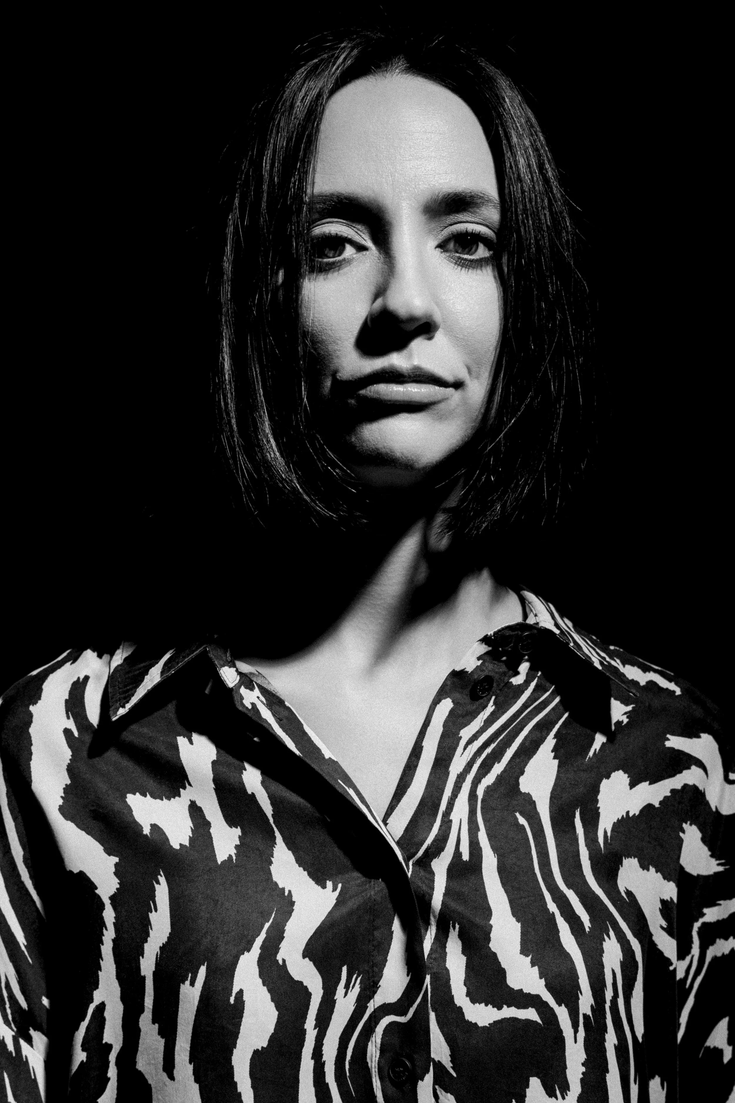Retrato em preto e branco de uma mulher usando uma camisa com estampa de zebra, iluminada dramaticamente contra um fundo escuro.