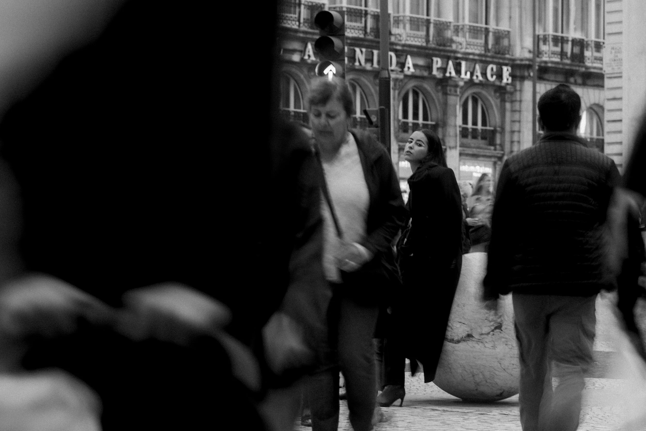 Cena urbana em preto e branco com pessoas caminhando; mulher no centro olhando para cima. Ao fundo, fachada do 'Avenida Palace'.
