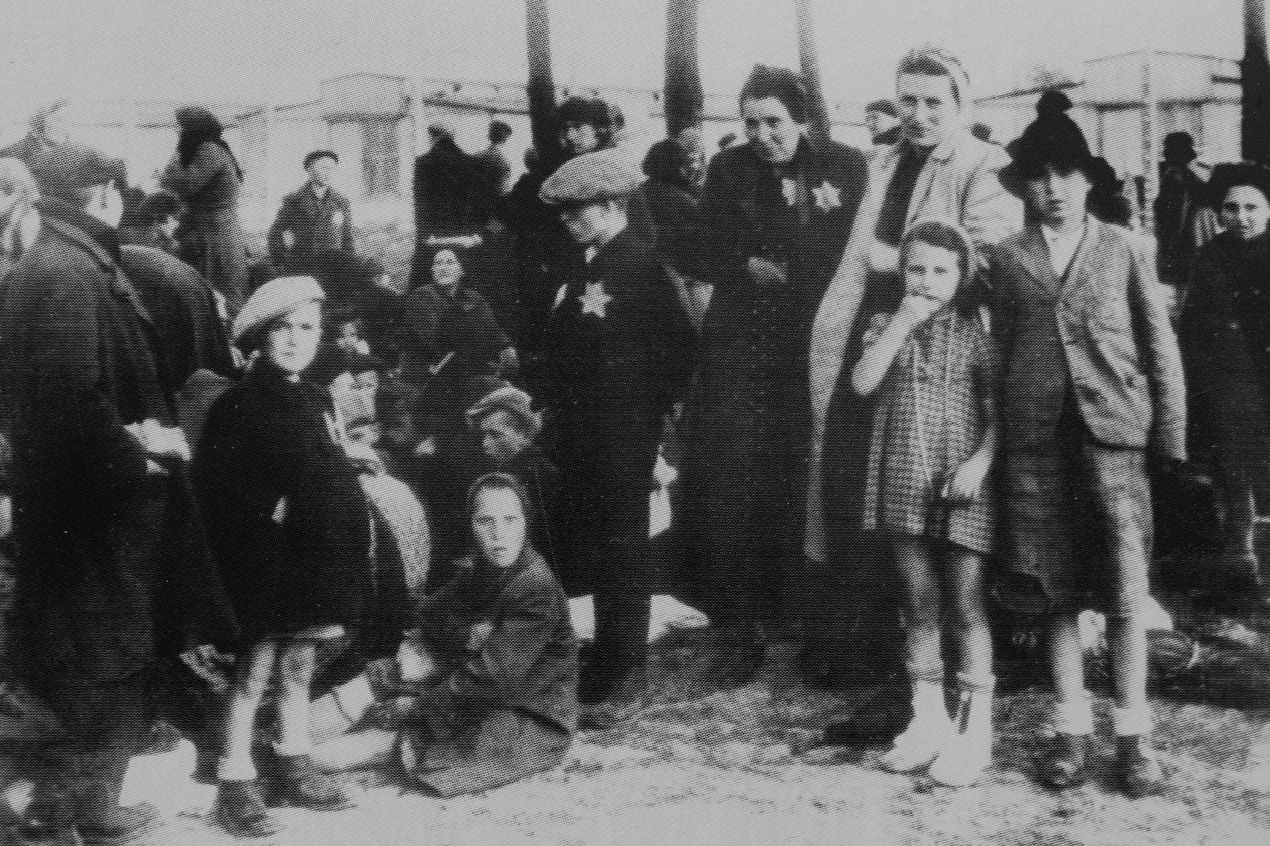 Grupo de pessoas, incluindo mulheres e crianças, usam estrelas de David, em um cenário de campo de concentração da Segunda Guerra Mundial. A imagem é em preto e branco.