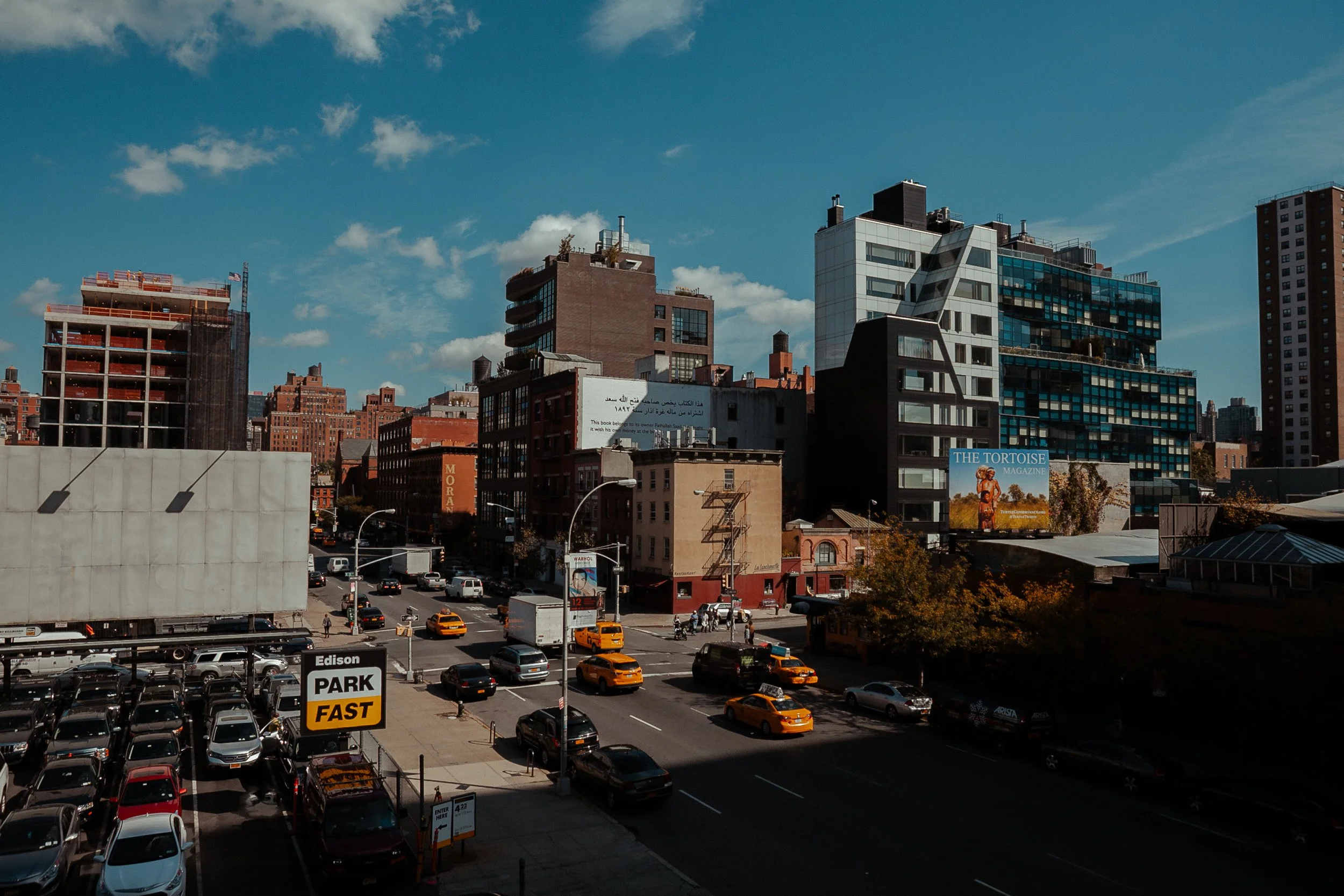 Vista urbana de uma cidade com prédios modernos e antigos, trânsito de táxis amarelos e carros, e um estacionamento ao ar livre. Há outdoors de propagandas nos edifícios.