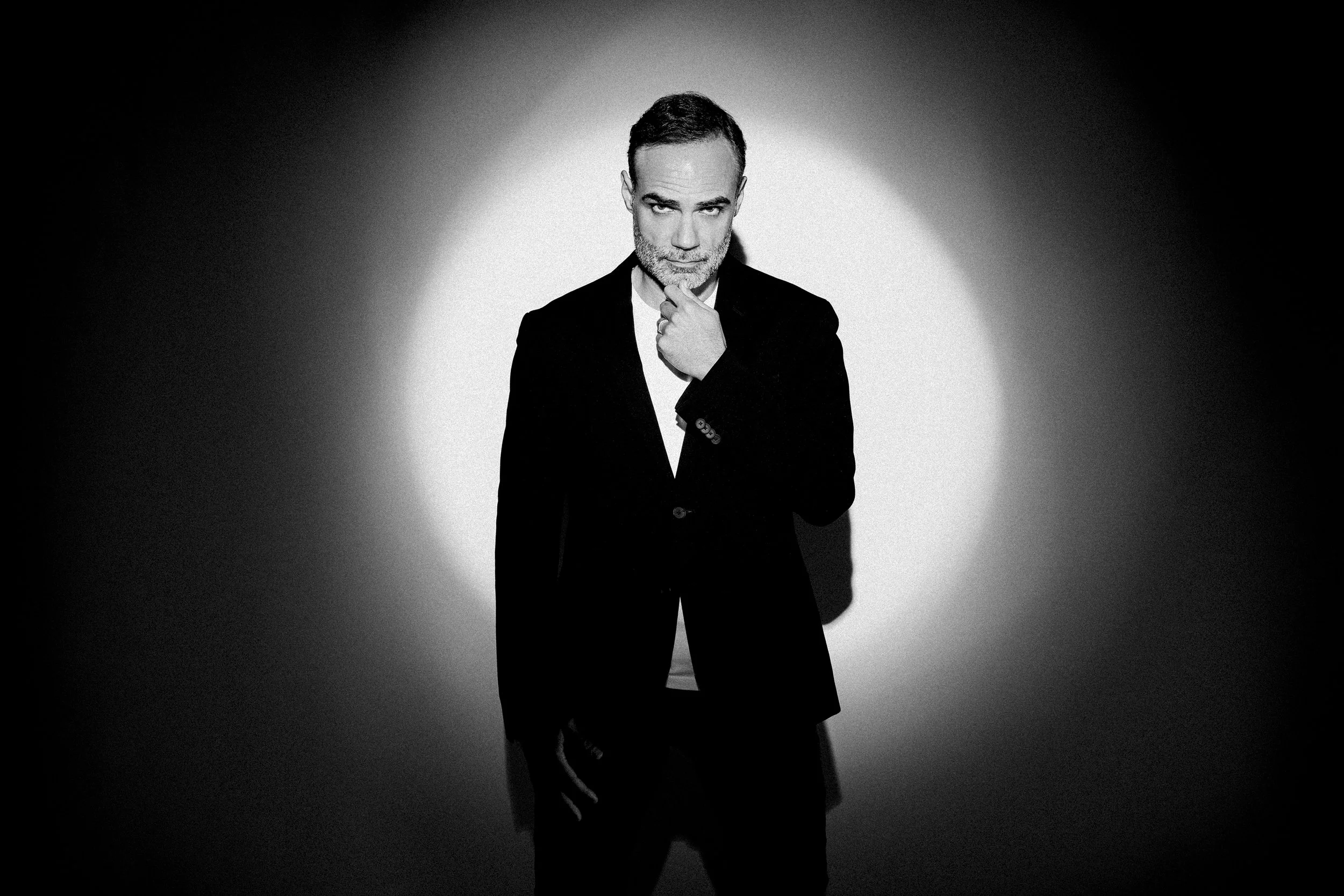 Homem em traje formal preto, posando em um fundo iluminado em preto e branco, com iluminação dramática. Ele tem expressão pensativa com a mão no queixo.