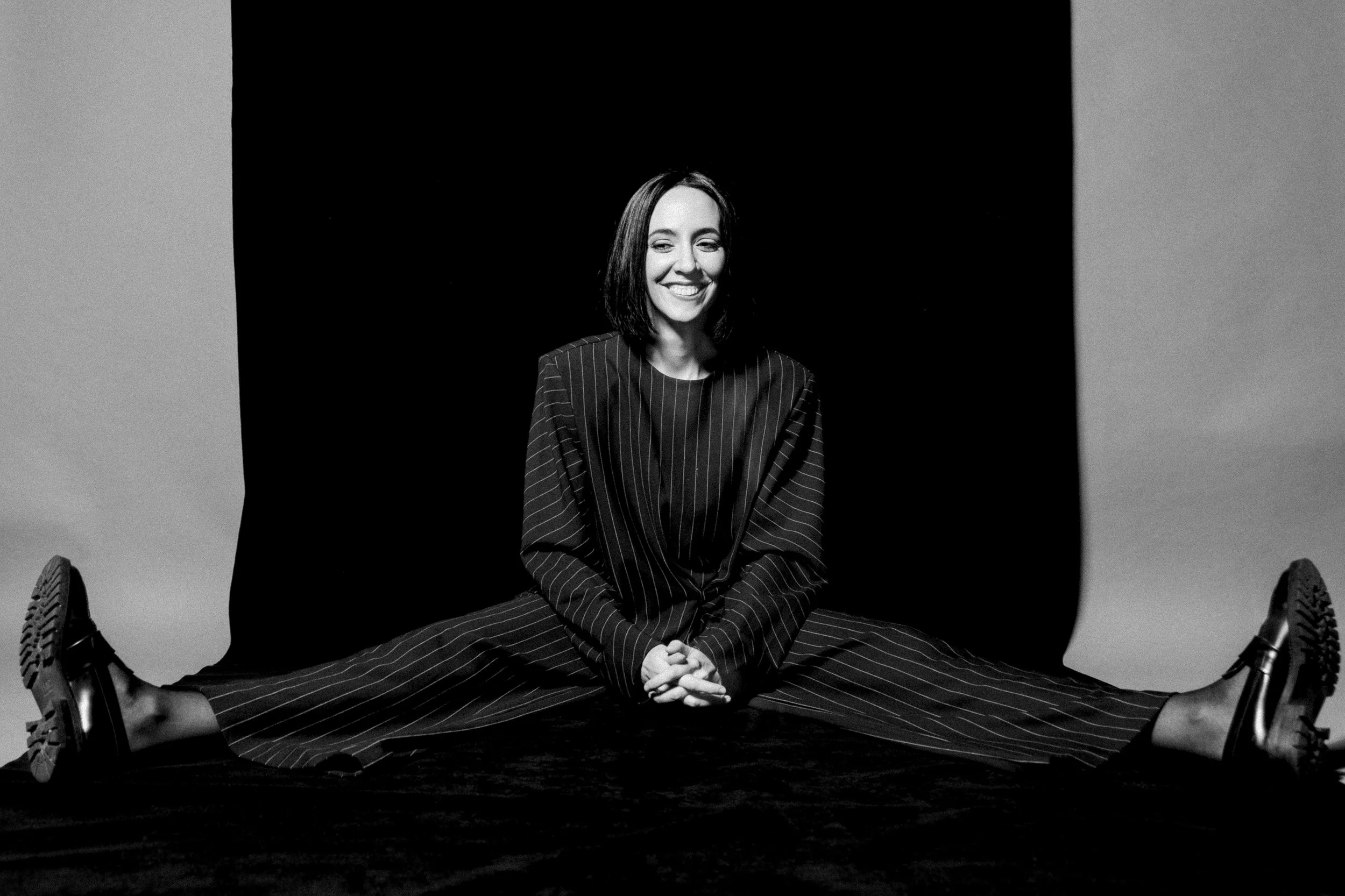 Pessoa sentada de pernas abertas, sorrindo, usando roupa formal com listras, em preto e branco.