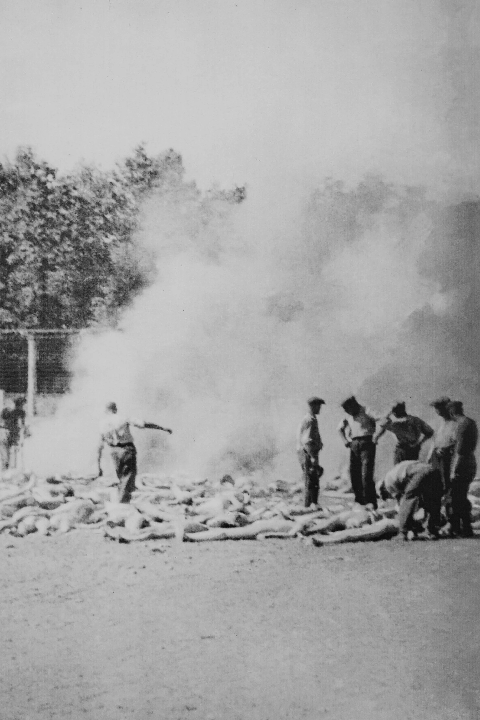 Imagem histórica em preto e branco mostrando um grupo de pessoas em torno de corpos no chão, com fumaça ao fundo, sugerindo um cenário de guerra ou conflito.