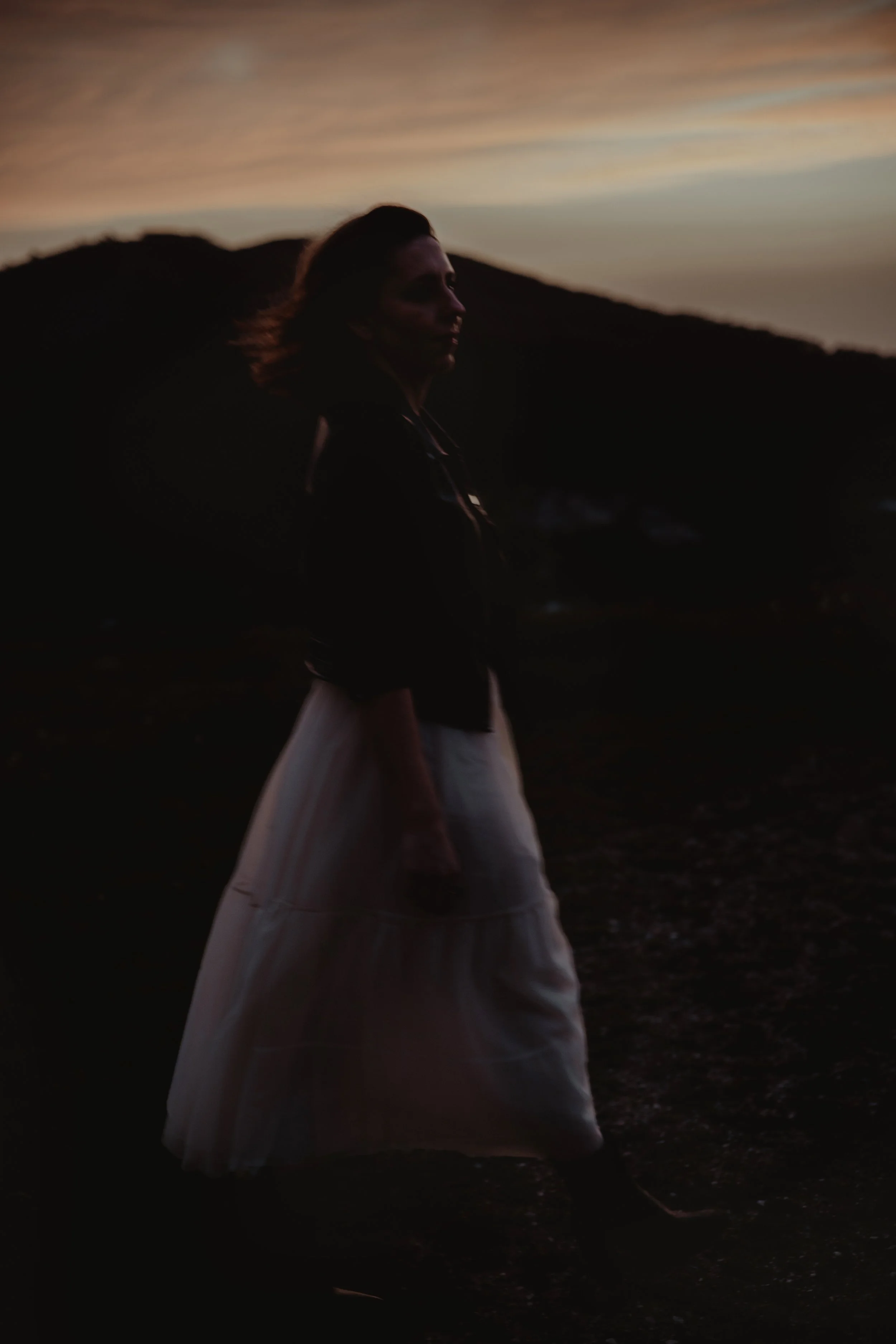 Pessoa com vestido claro e jaqueta escura ao pôr do sol, em um cenário de montanhas.