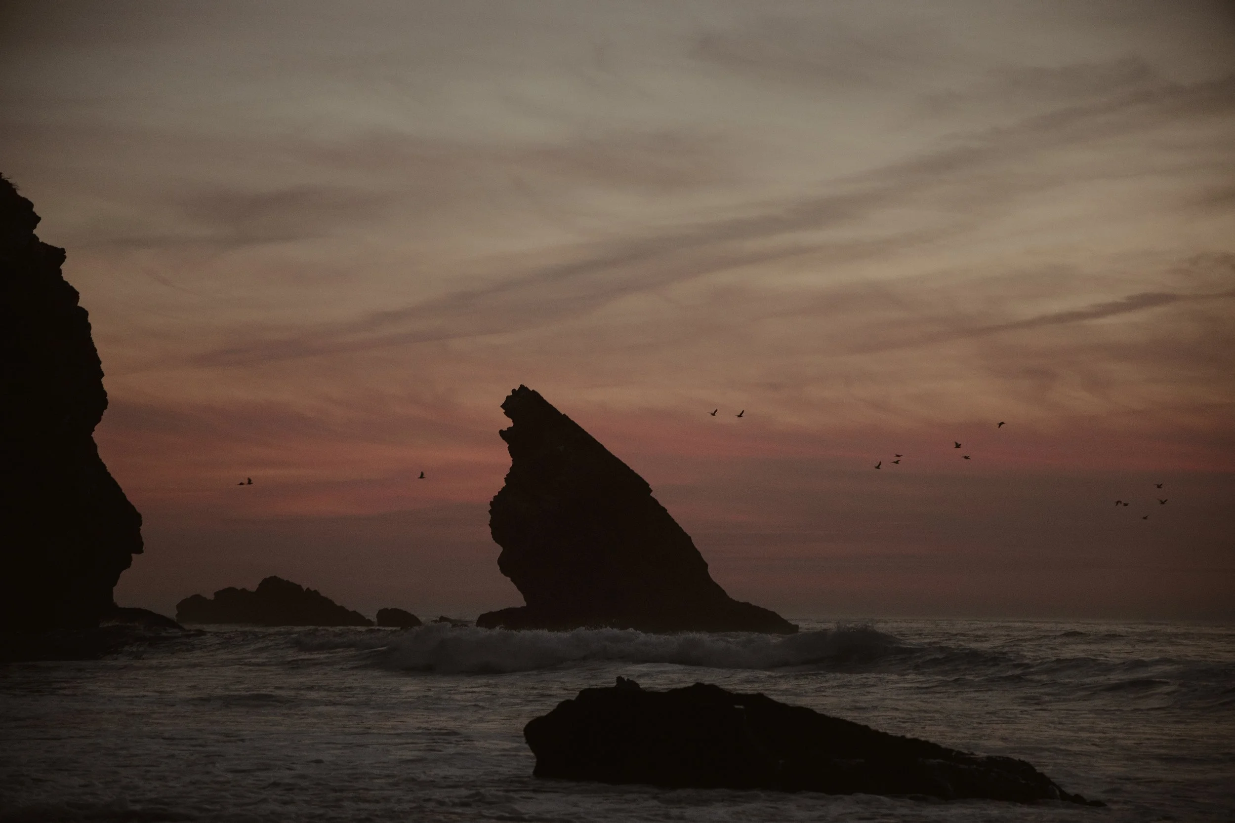Formações rochosas no mar ao entardecer com pássaros voando.