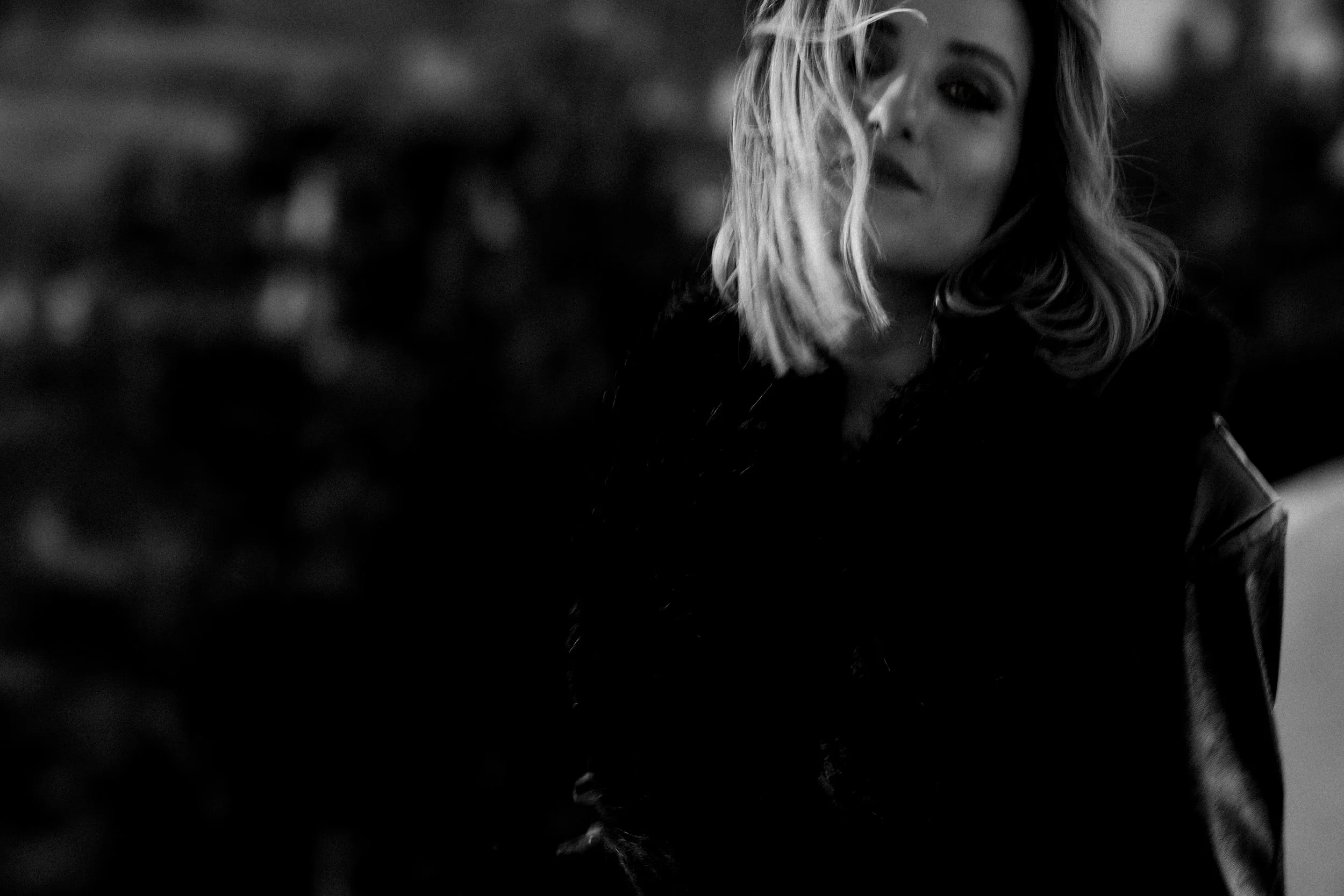 Mulher loira em fotografia em preto e branco com cabelo ao vento, usando casaco de pele, fundo desfocado.