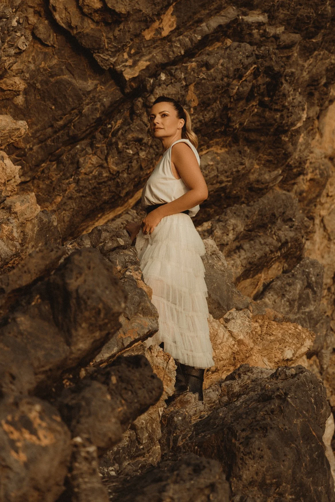 Mulher em vestido branco posando entre rochas marrons, olhar distante, cenário natural. Moda e natureza combinados.